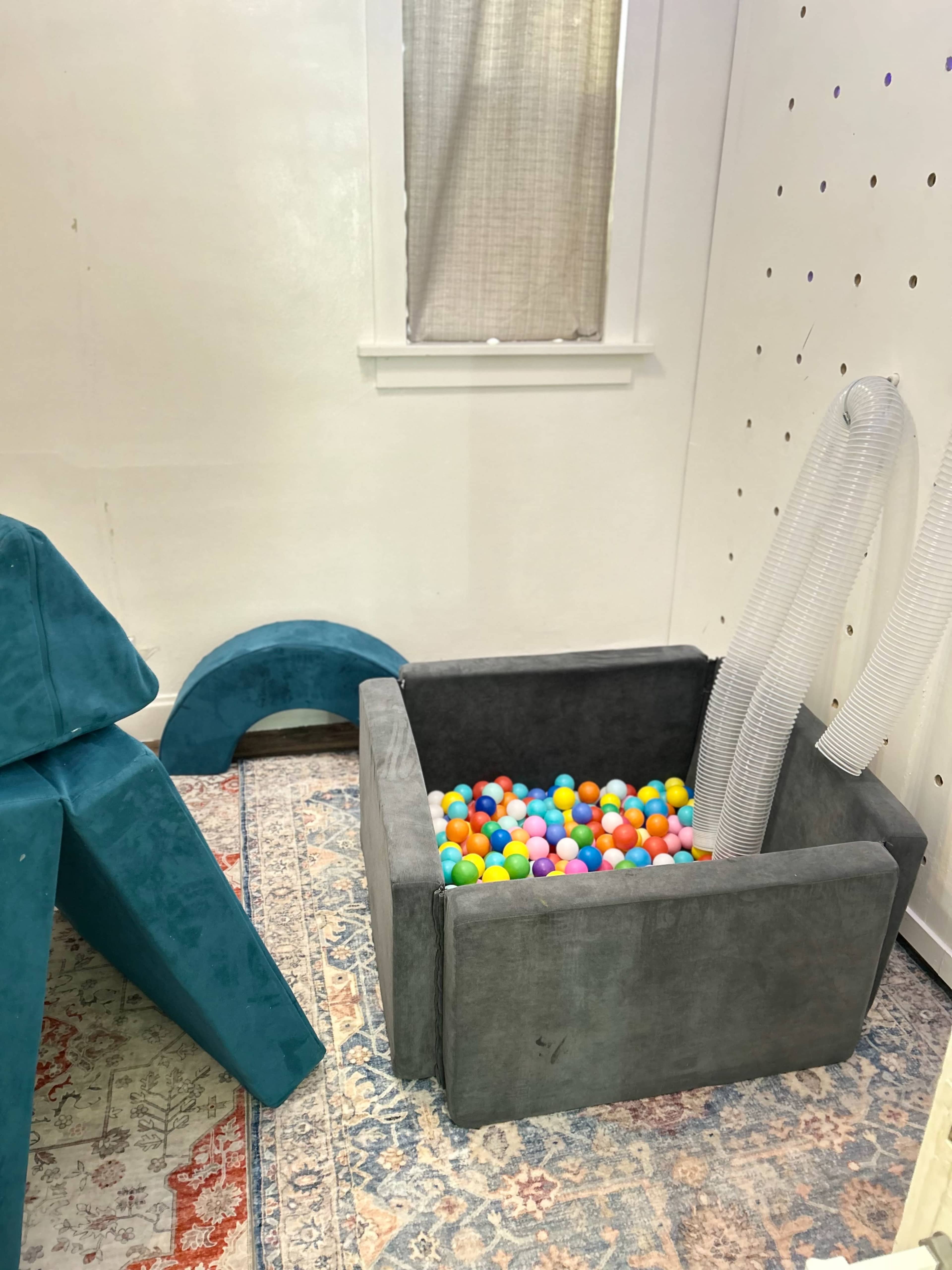 The image shows a playroom with a gray ball pit filled with colorful balls, soft teal shapes for climbing, and a patterned rug on the floor.