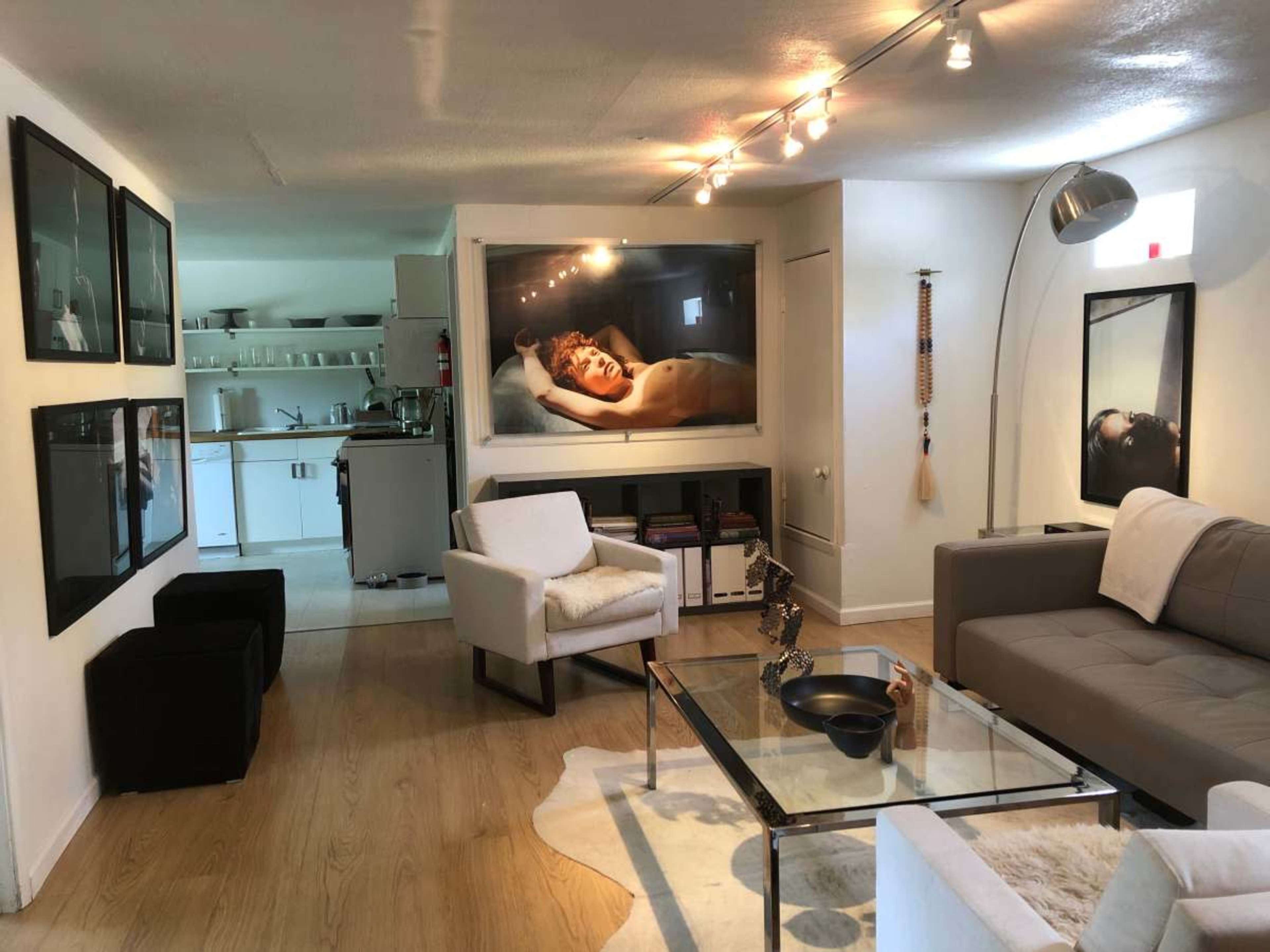 The image shows a modern living space with a kitchen in the background, featuring a glass coffee table, a beige couch, and several framed photographs on the walls.