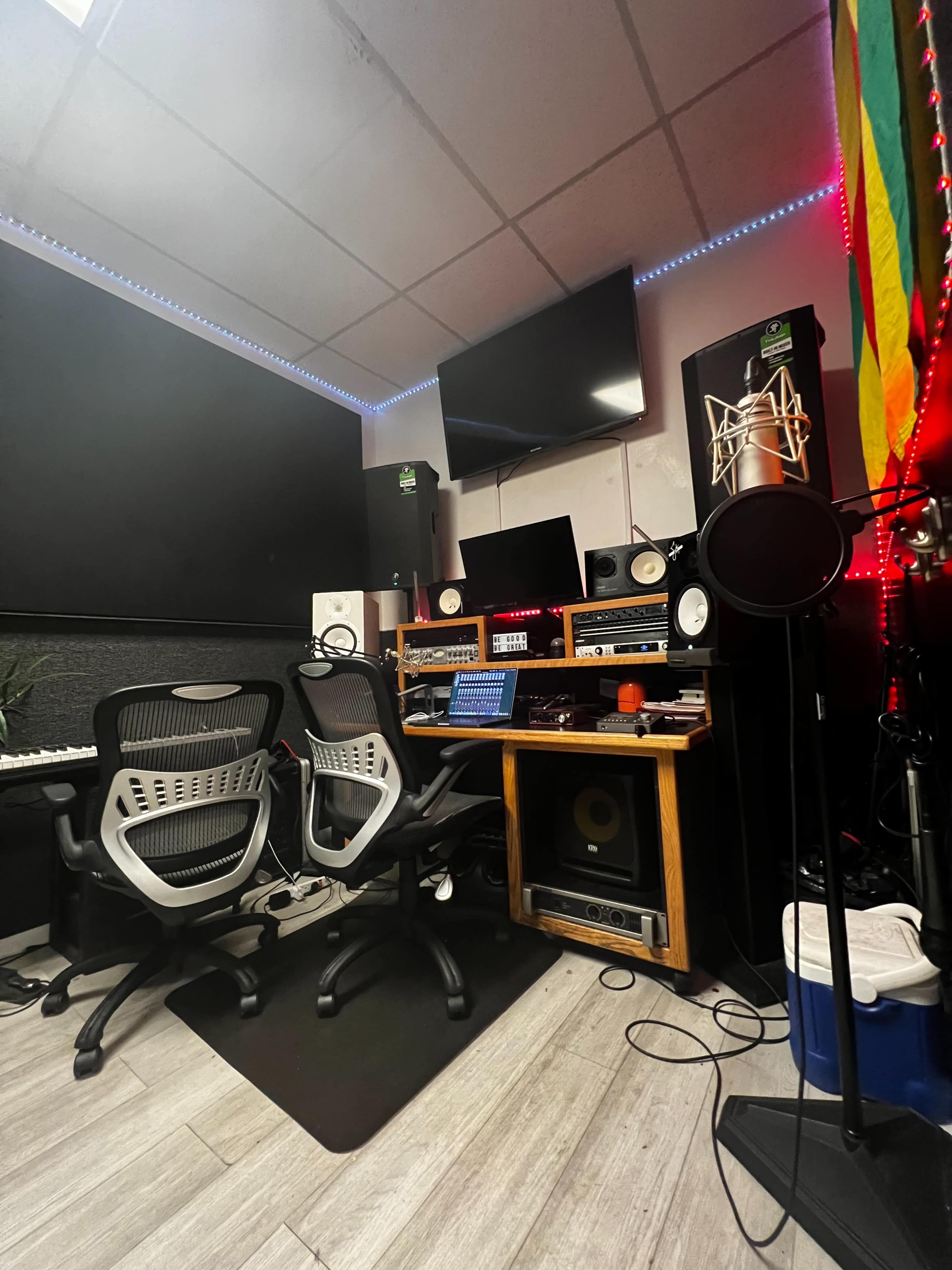 A music recording studio setup with dual chairs, audio equipment, a microphone stand, and a television mounted on the wall.