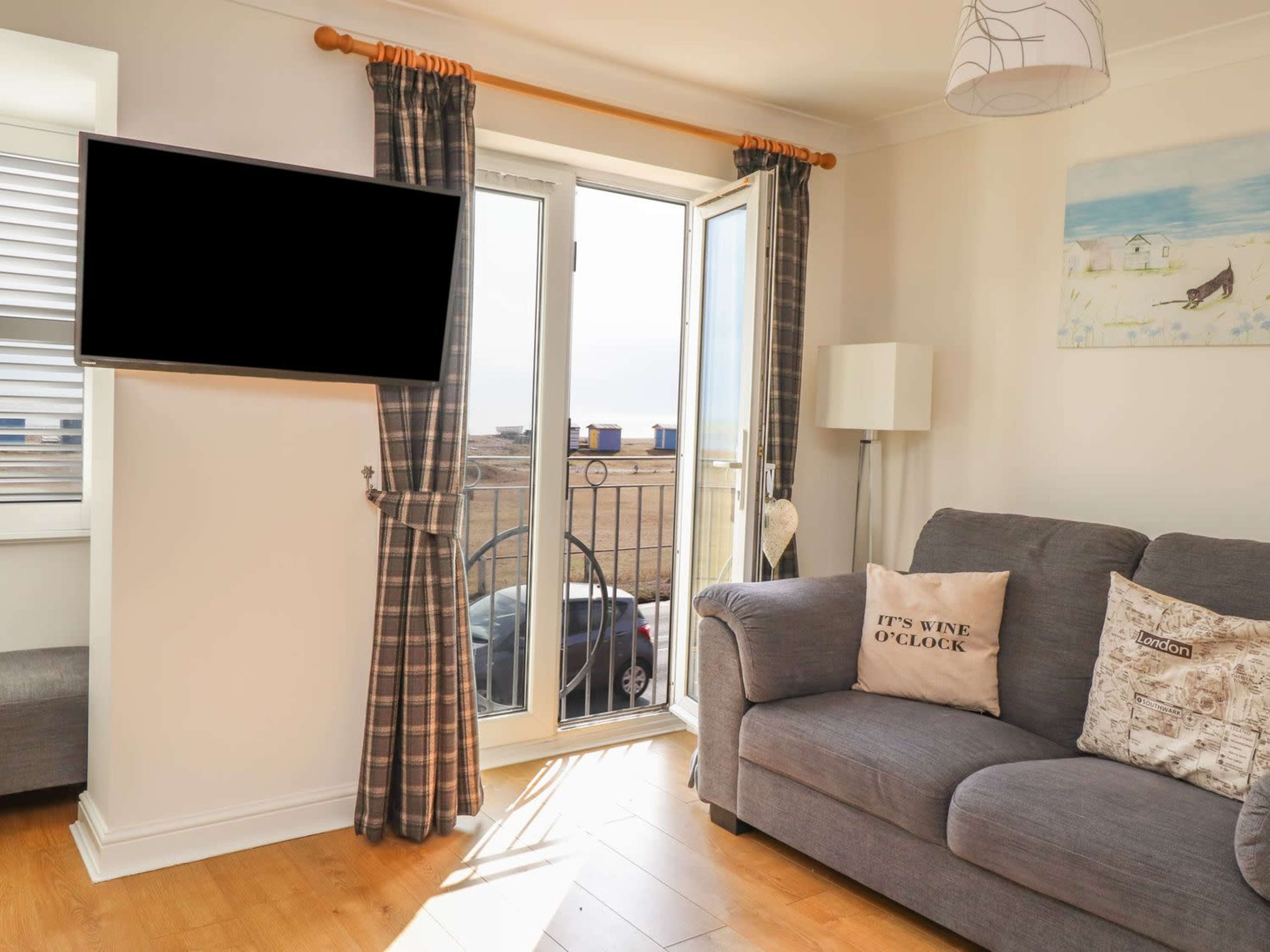 A living room features a gray sofa with decorative pillows, a wall-mounted television, and a balcony door that opens to an outdoor view.
