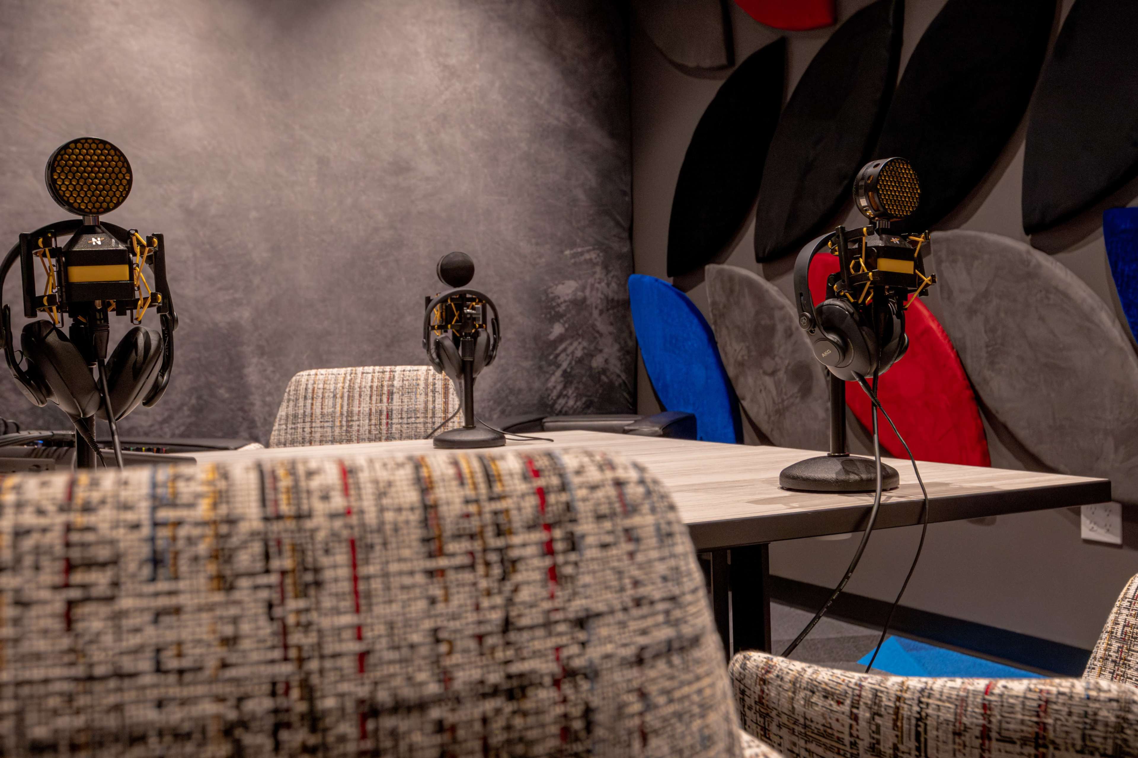 The image shows a podcast studio with two microphones, headphones on stands, and a table surrounded by textured chairs, set against a colorful wall.
