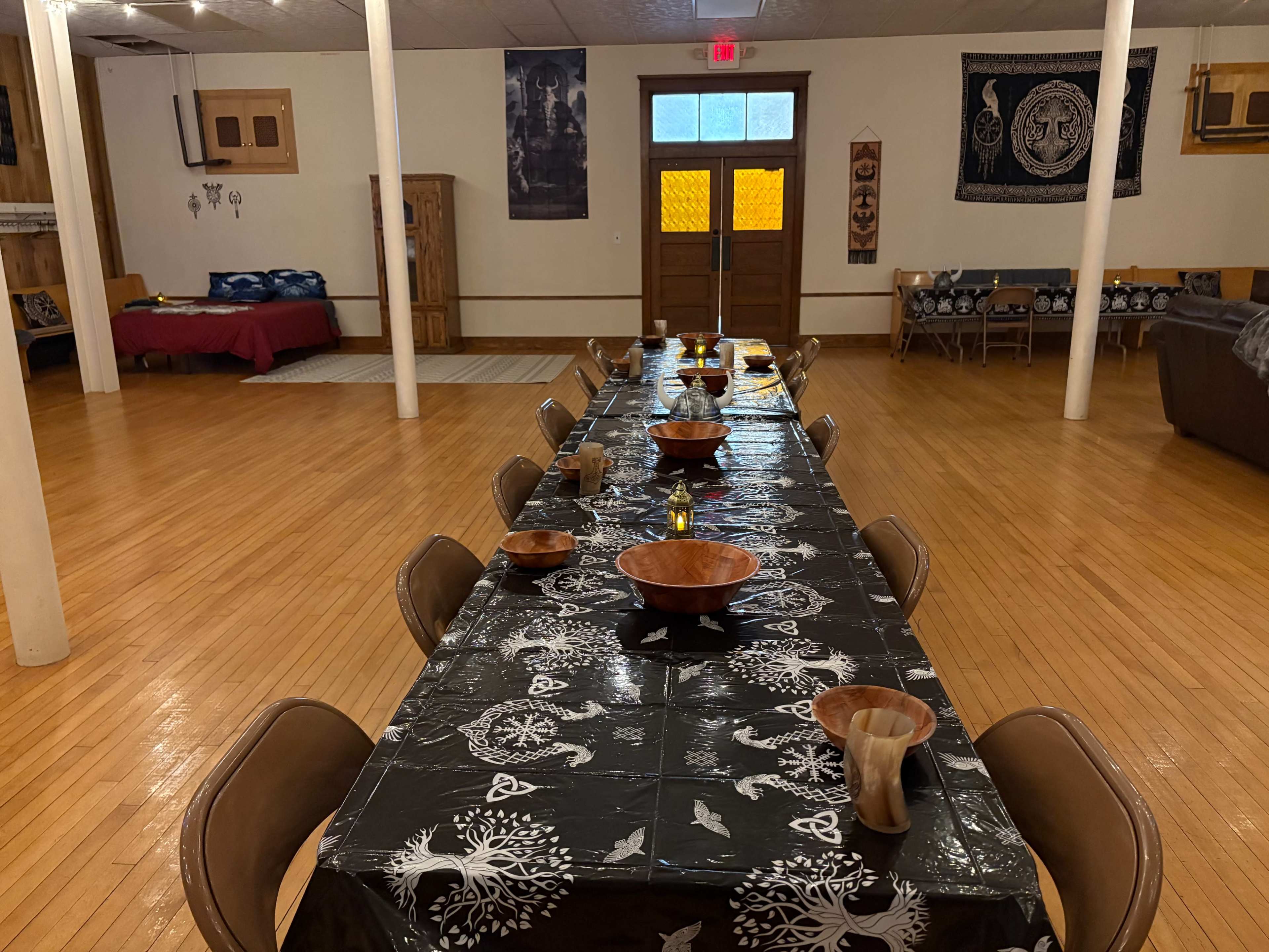 A long dining table is set with wooden bowls and black tablecloths, surrounded by chairs in a spacious, well-lit room with wooden floors and decorative wall hangings.