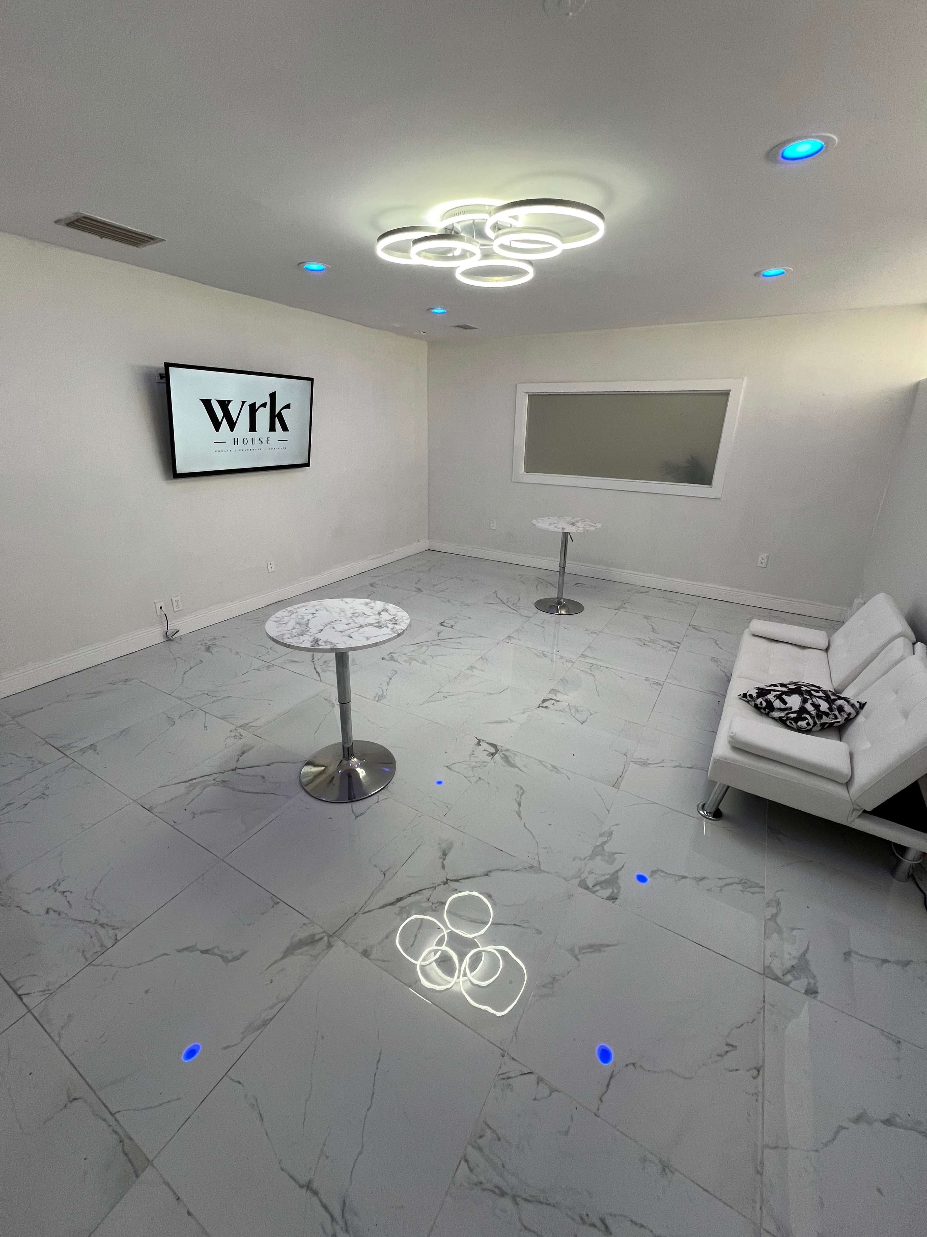 The image features a modern, minimalist interior space with marble flooring, a white sofa, two small tables, and a large screen displaying the word "Wrk."