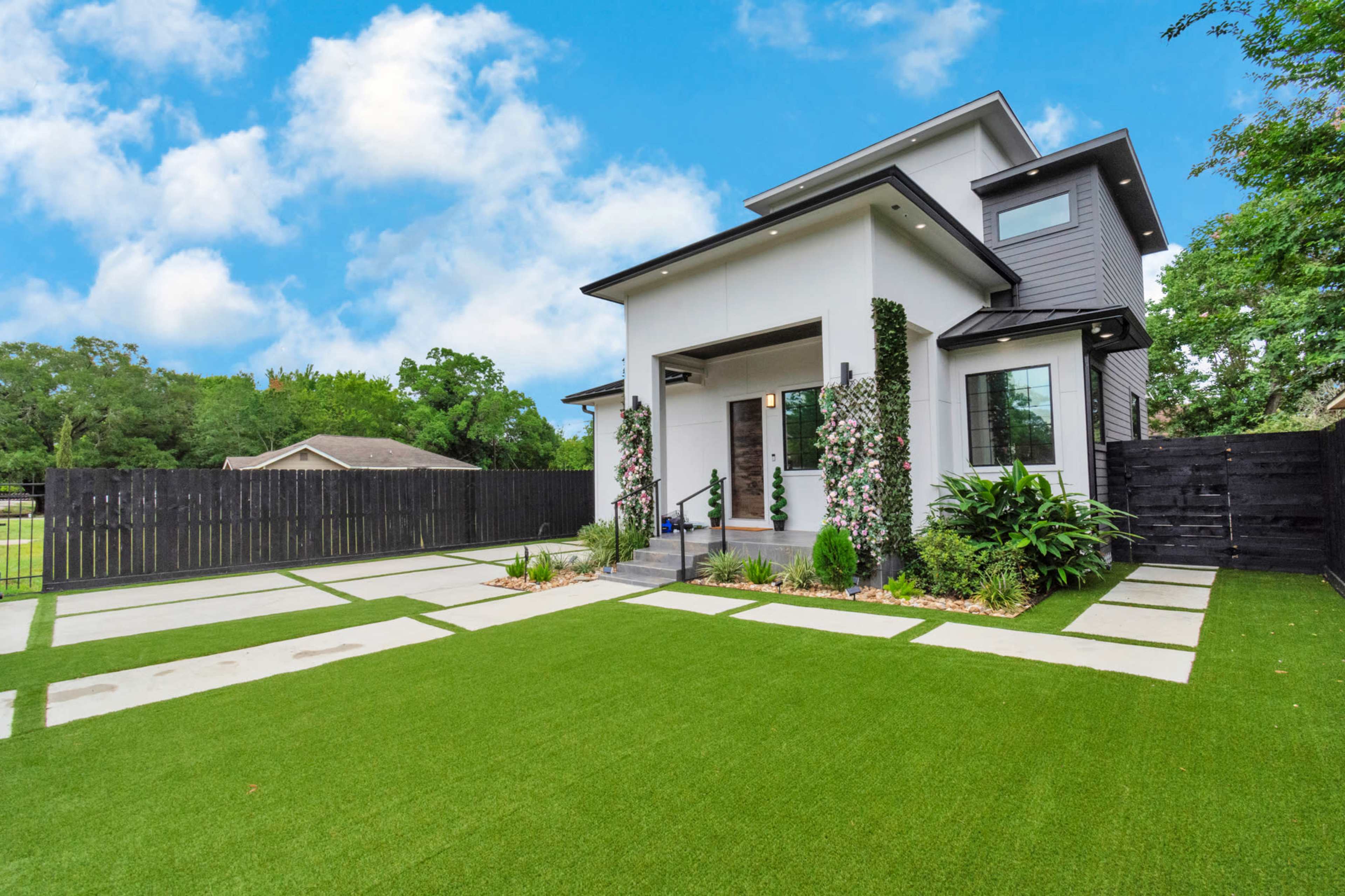 A modern two-story house with a landscaped front yard featuring artificial turf and concrete pathways, bordered by a black wooden fence.