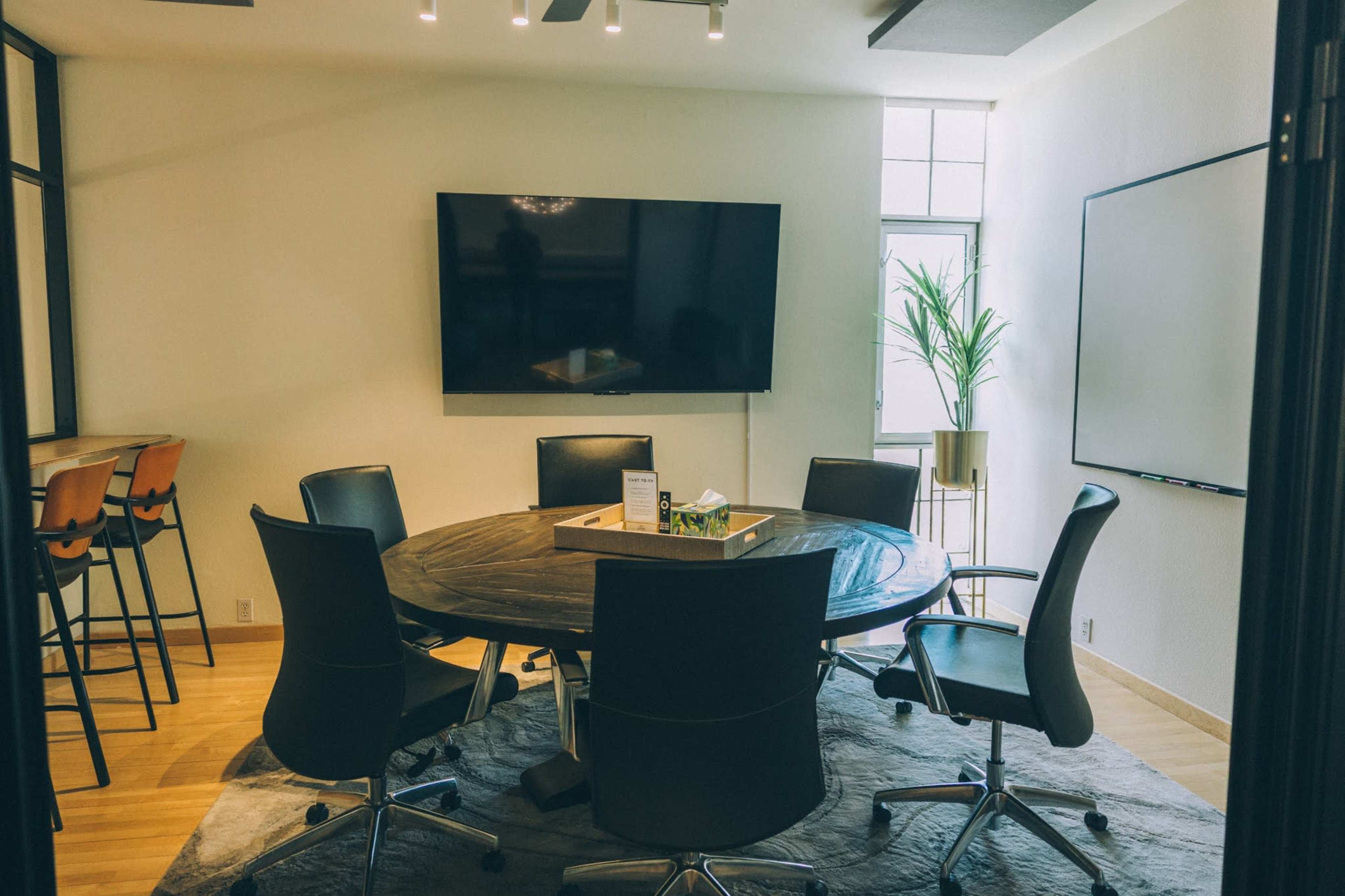 The image shows a modern conference room with a round wooden table, six black chairs, a large wall-mounted TV, and a whiteboard.