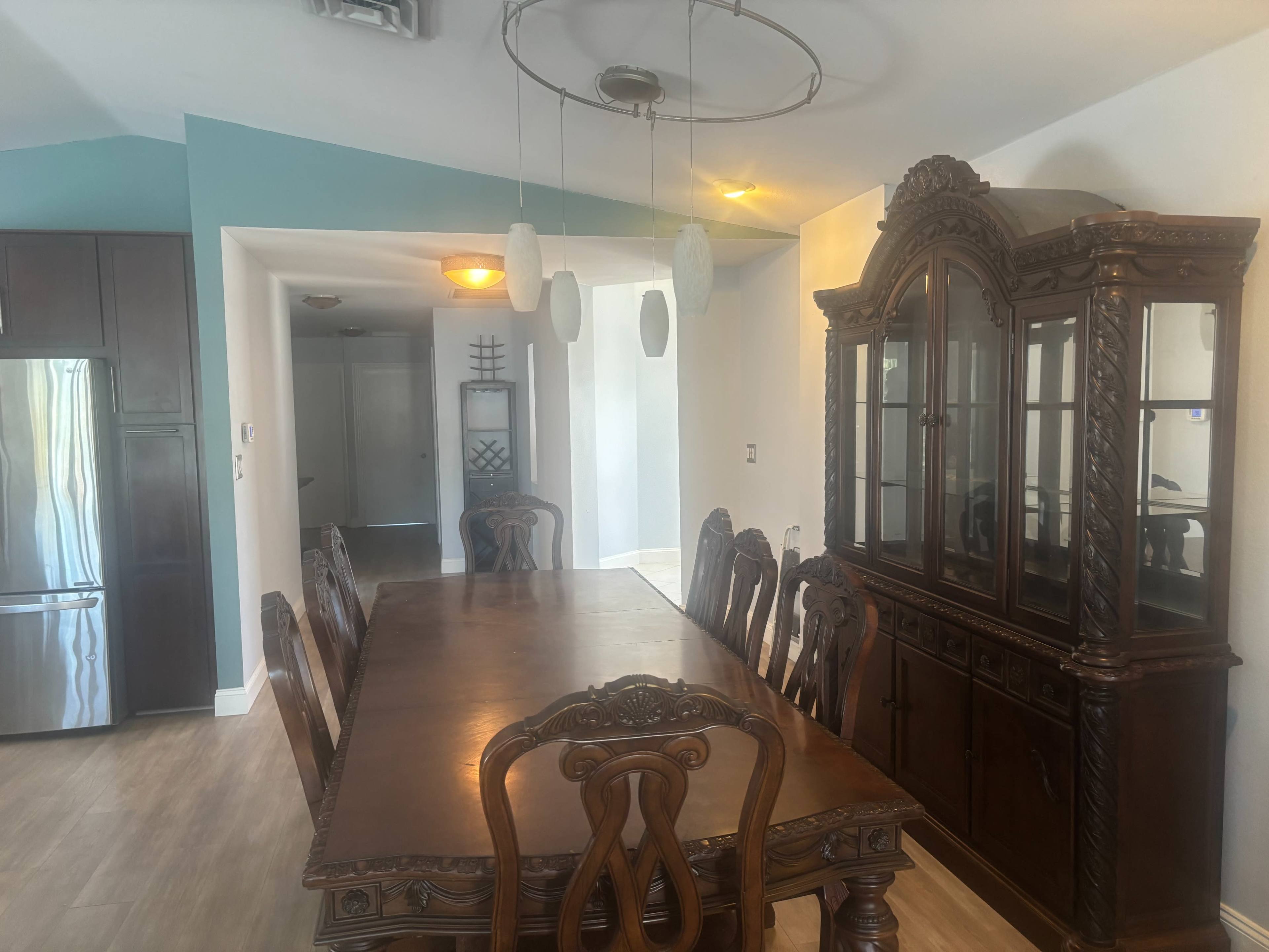 A dining room features a large wooden table surrounded by ornate chairs and a glass-fronted china cabinet, with modern pendant lights overhead.