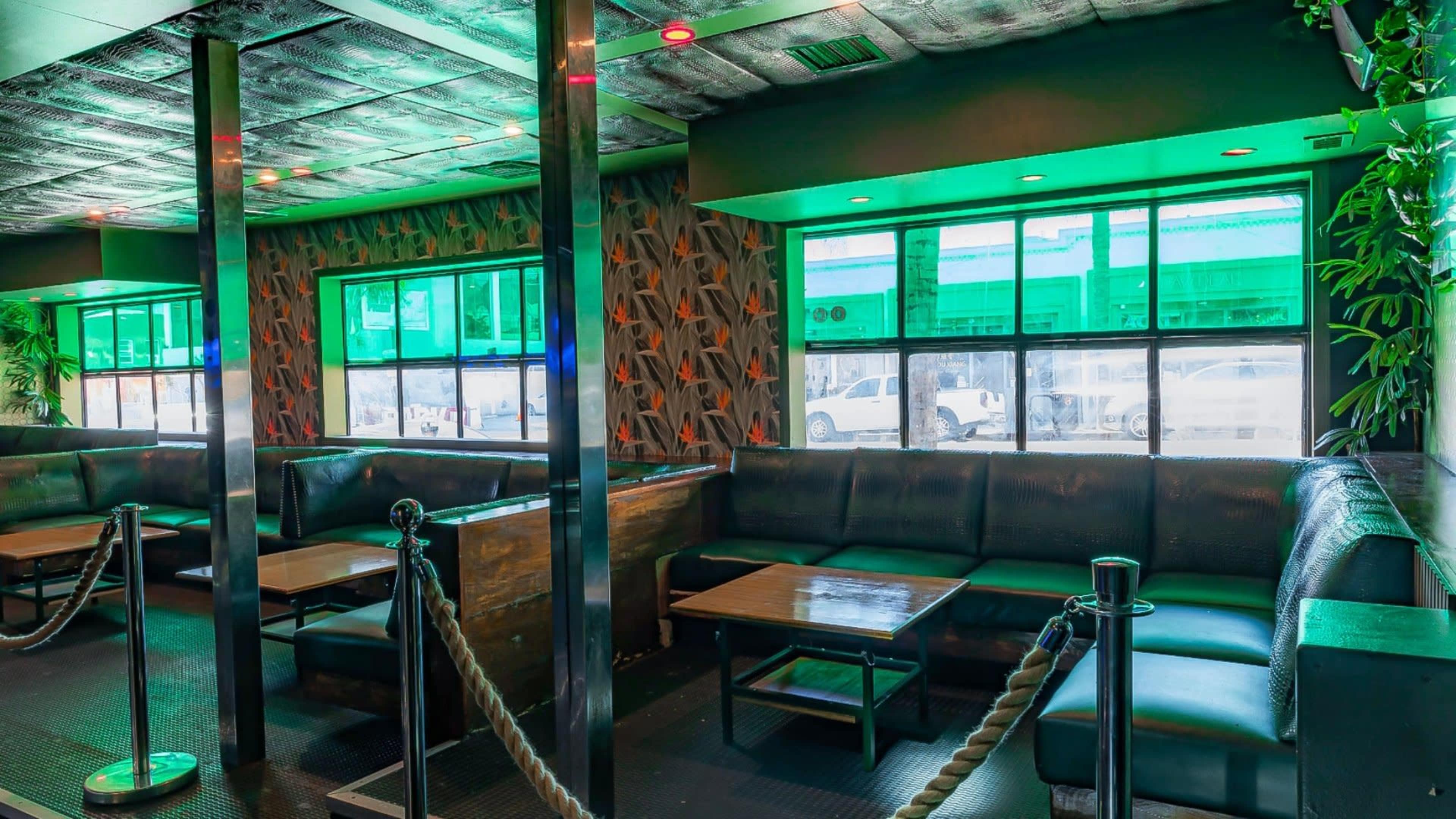 The interior of a lounge features dark leather seating, wooden tables, and large windows illuminated by green lights.