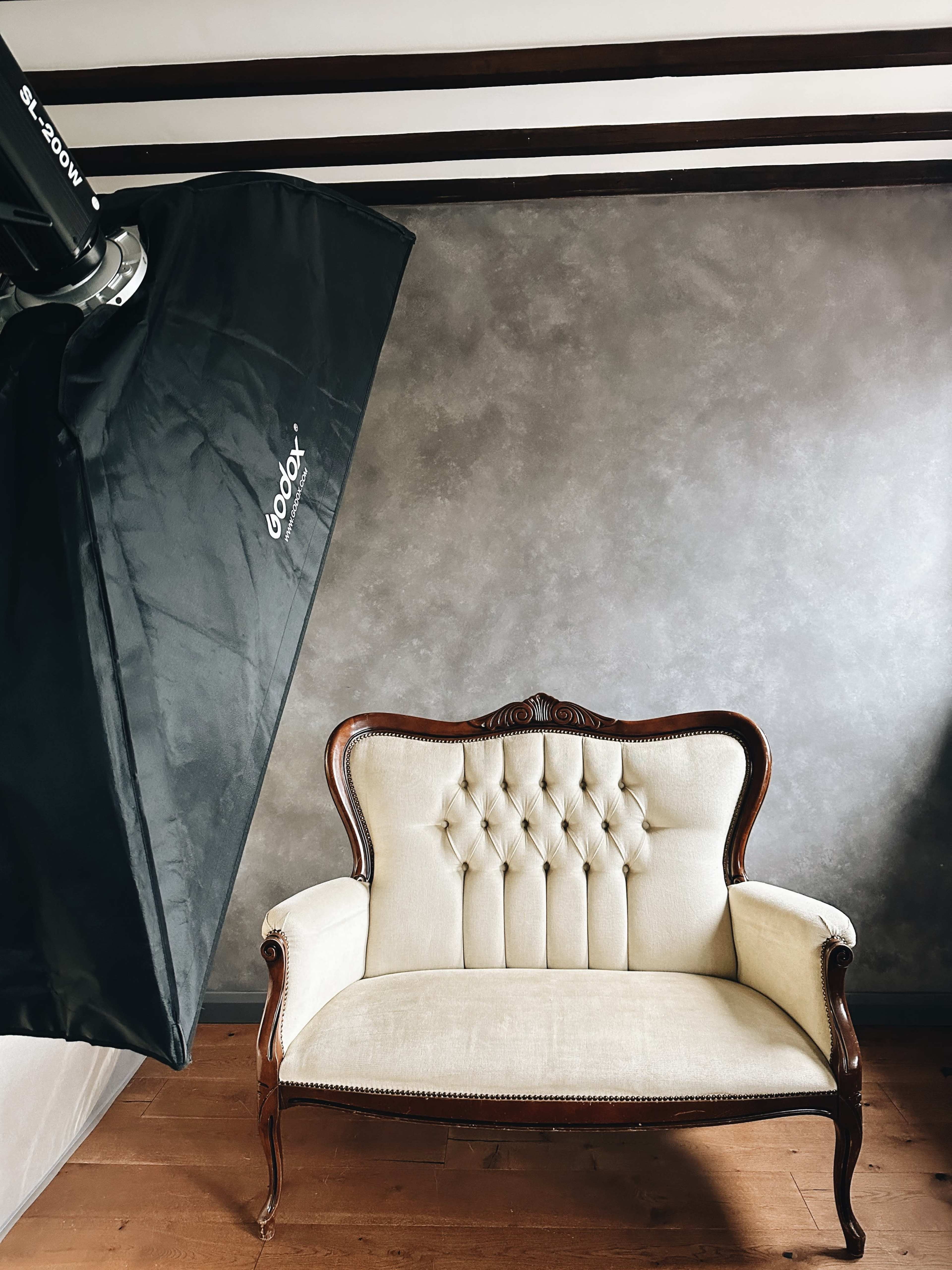 A vintage light-colored loveseat is positioned in front of a gray textured wall, with a softbox light setup nearby.