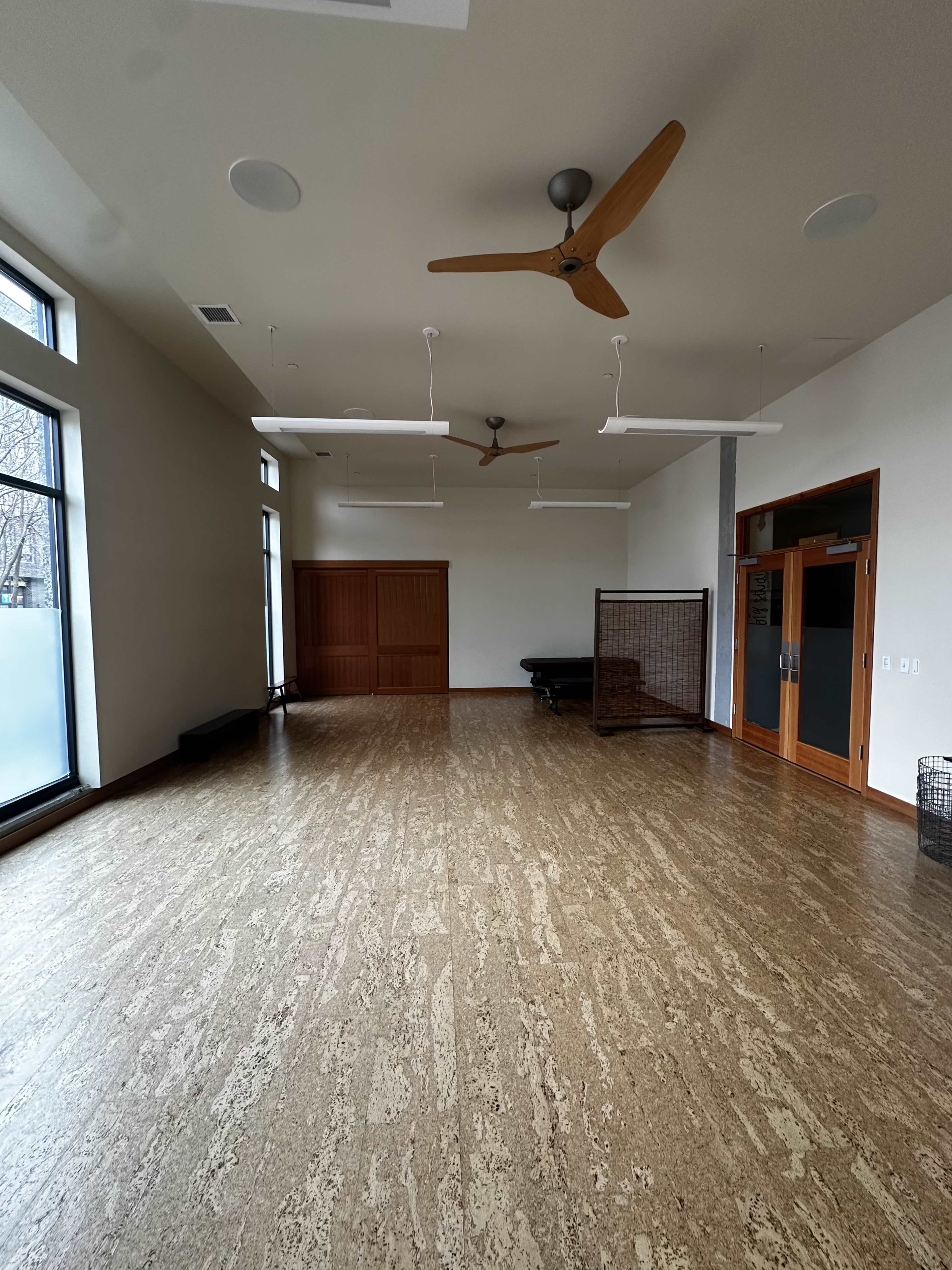 A spacious, empty room with large windows, wooden ceiling fans, and a wooden sliding door on one side.