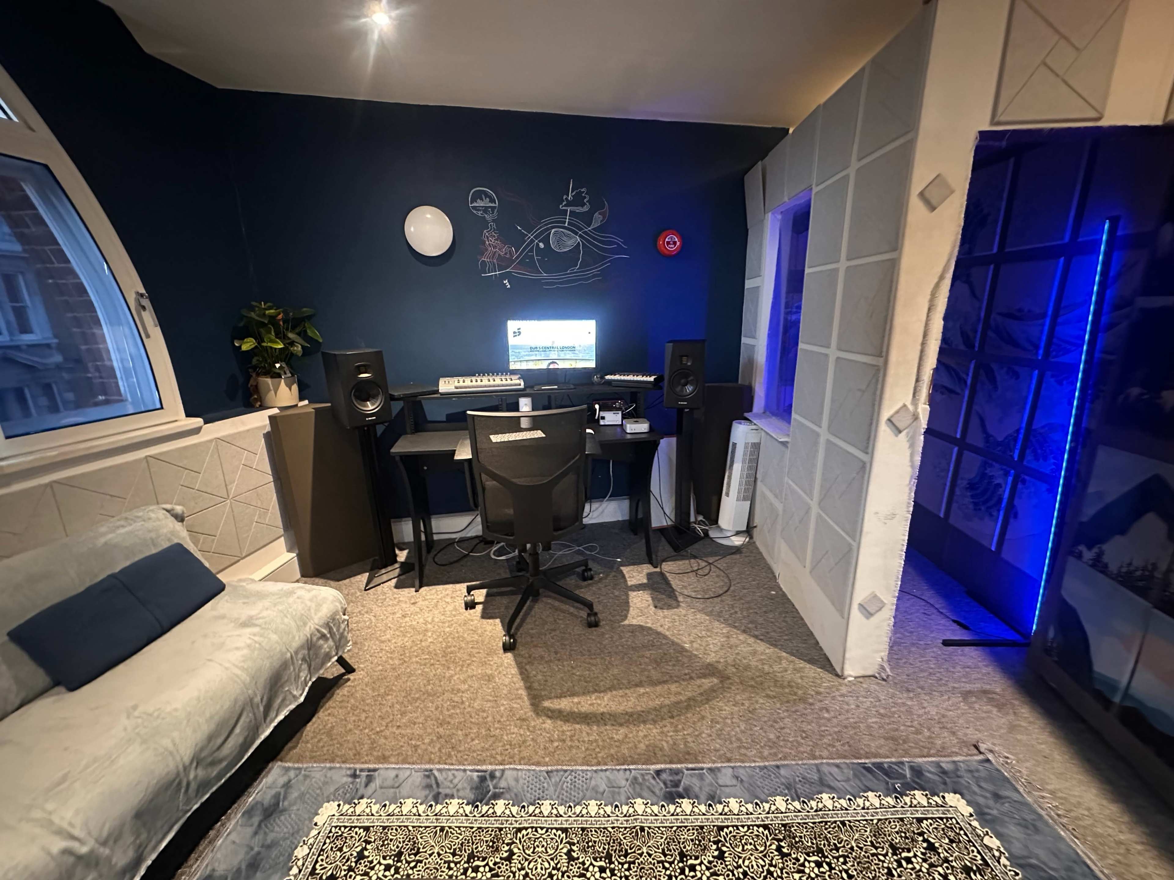 The image shows a small, cozy recording studio with a computer desk, audio speakers, and a couch, all set against a blue wall.