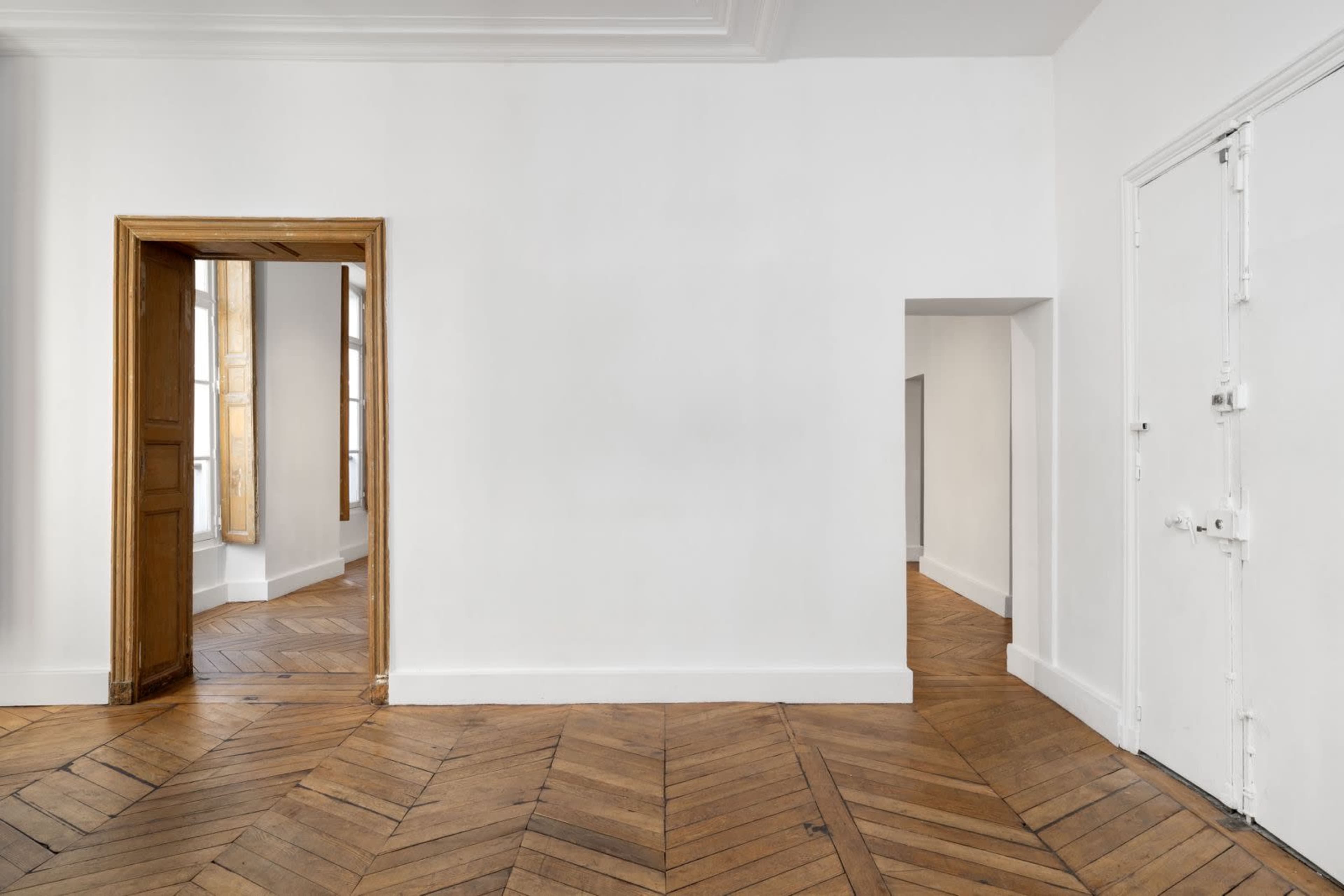 The image shows a minimalist room with white walls, wooden herringbone flooring, and two doorways leading to other spaces.