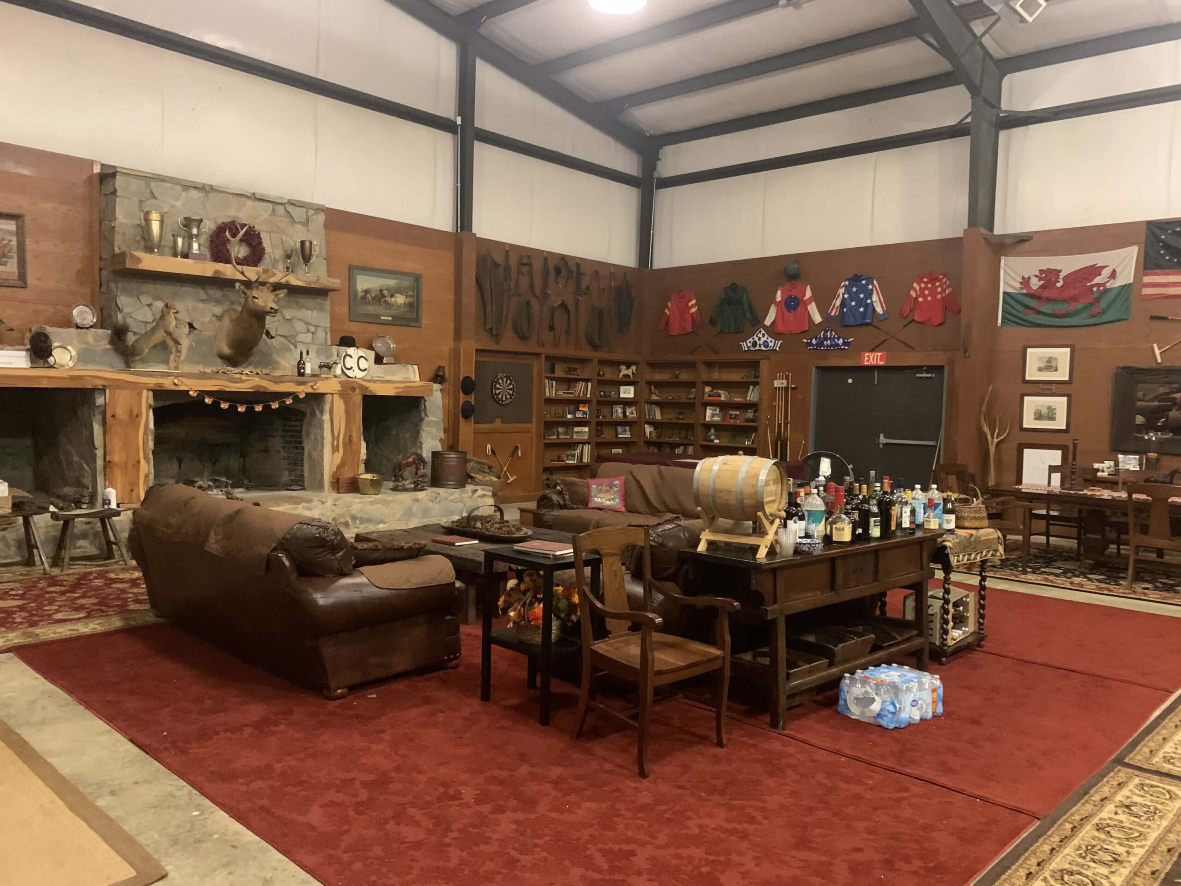 The image shows a spacious rustic room with a seating area featuring leather sofas, a large stone fireplace, and various decorations, including mounted animal heads and shelves filled with bottles and memorabilia.