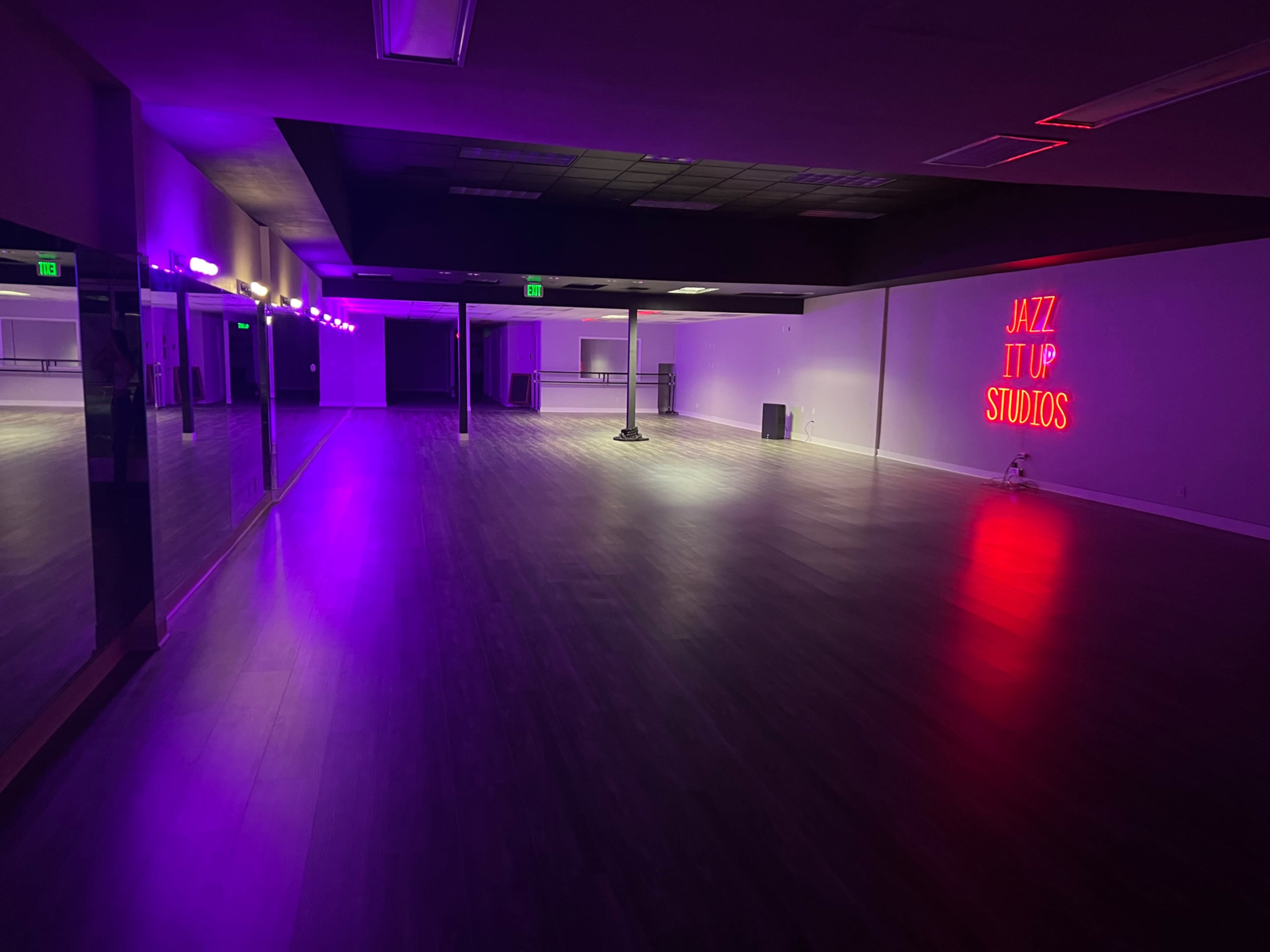 The image shows a spacious dance studio with purple and neon lighting, featuring a mirrored wall and a sign that reads "JAZZ IT UP STUDIOS."
