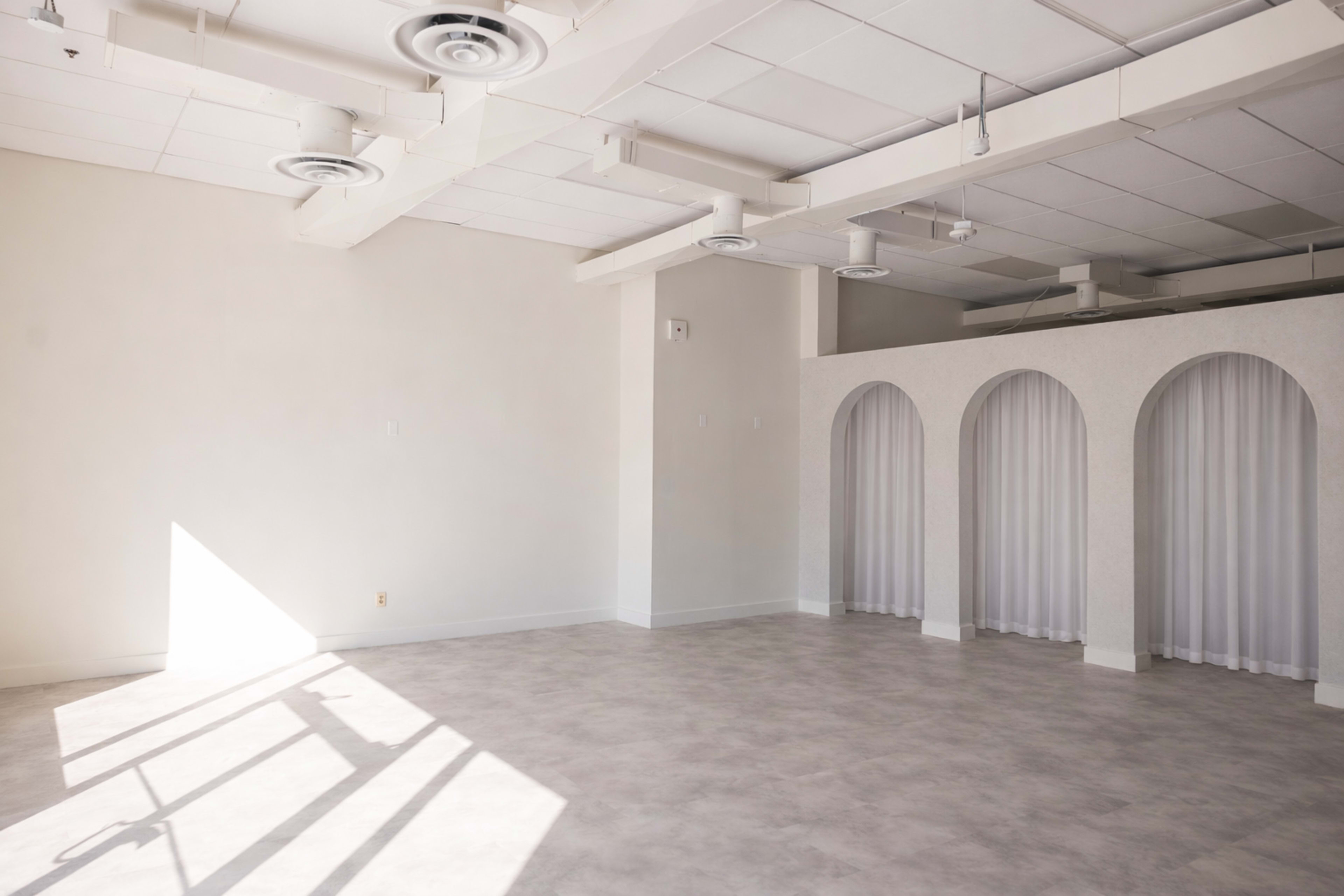 The image shows a spacious, empty room with light-colored walls, a smooth floor, and architectural arches with curtains on one side.