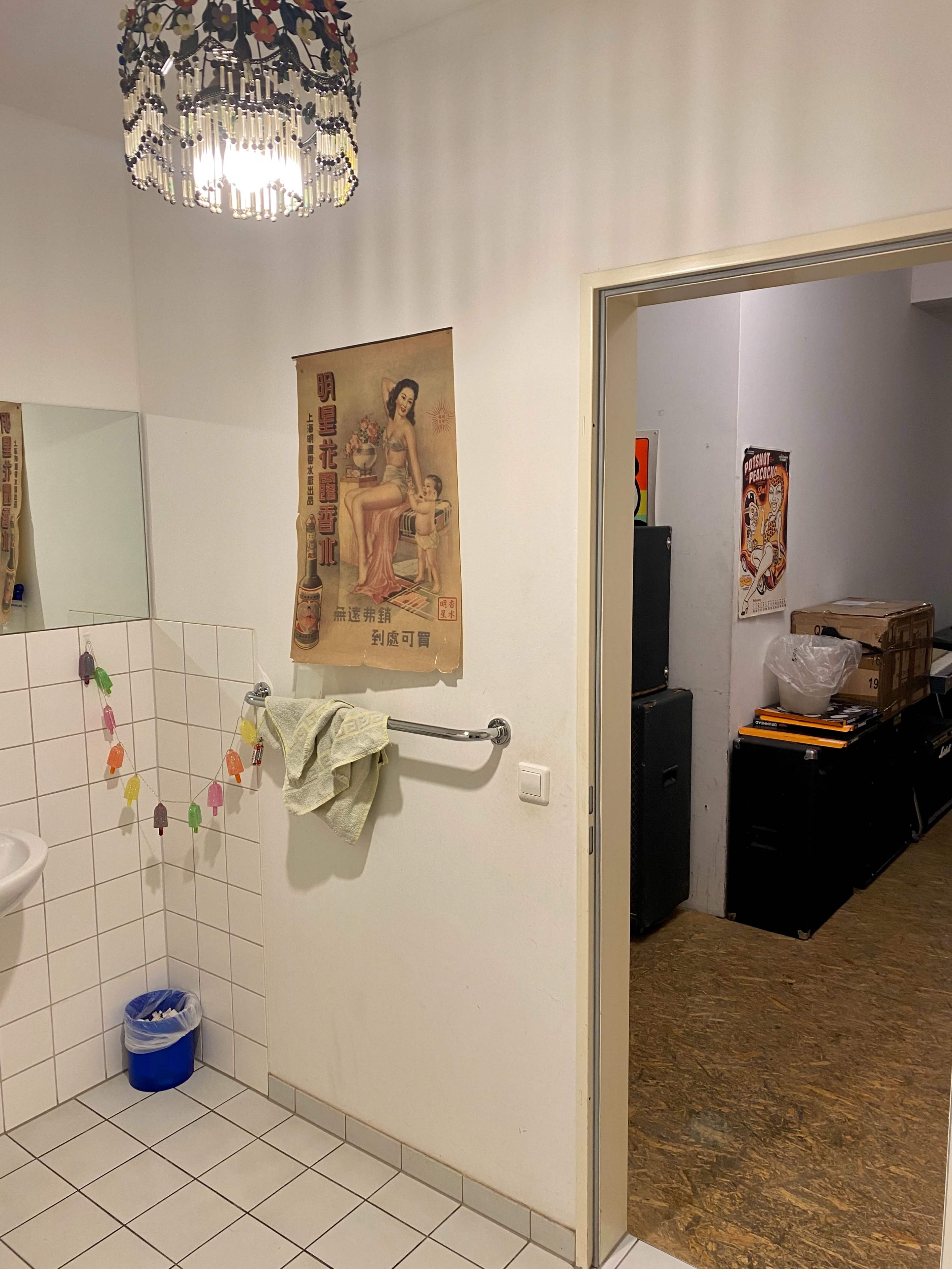 A bathroom with a vintage poster on the wall, a mirror, and a doorway leading to a cluttered room.