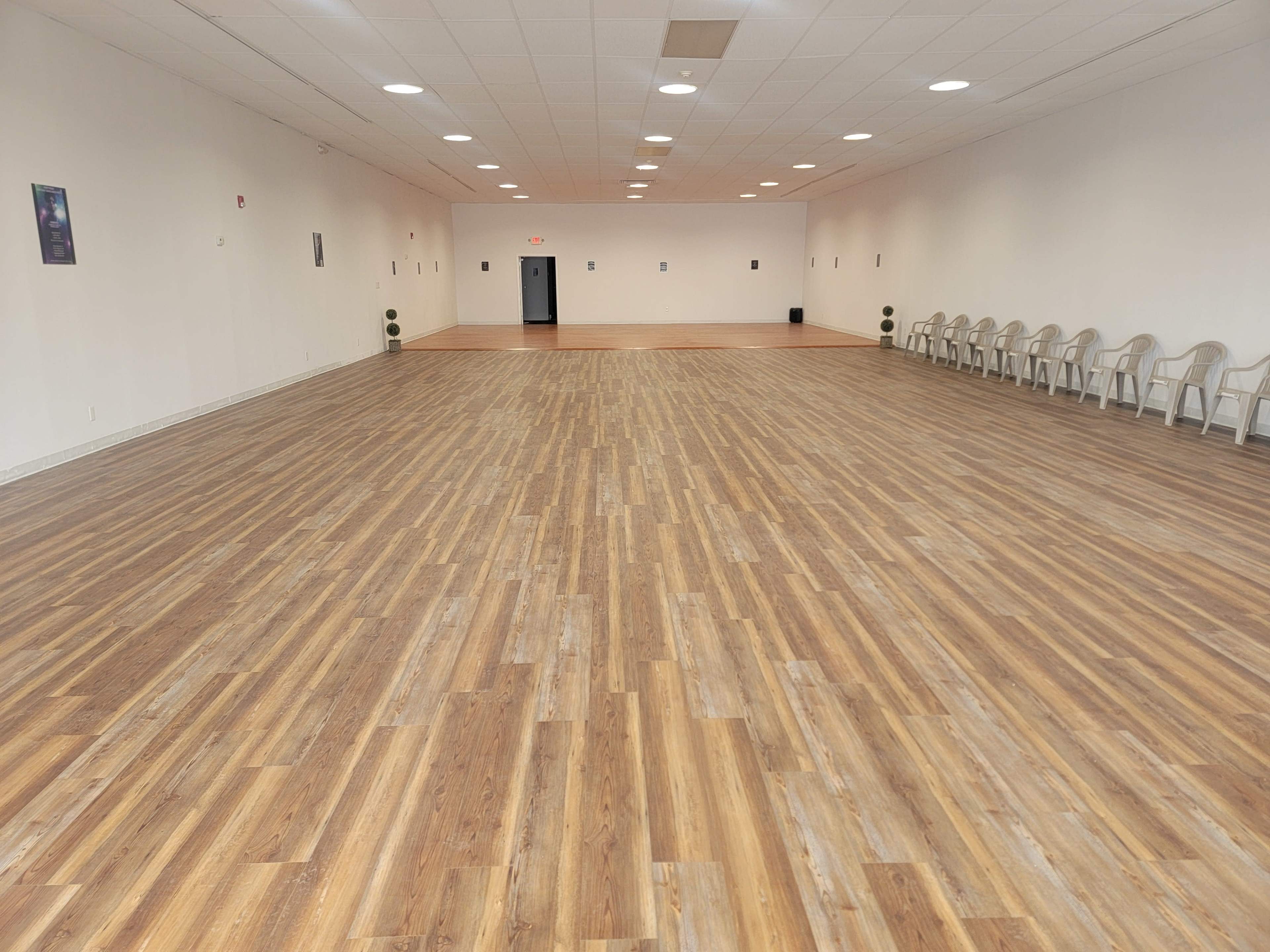 A spacious room with wooden flooring, featuring a row of chairs along one side and plain white walls.