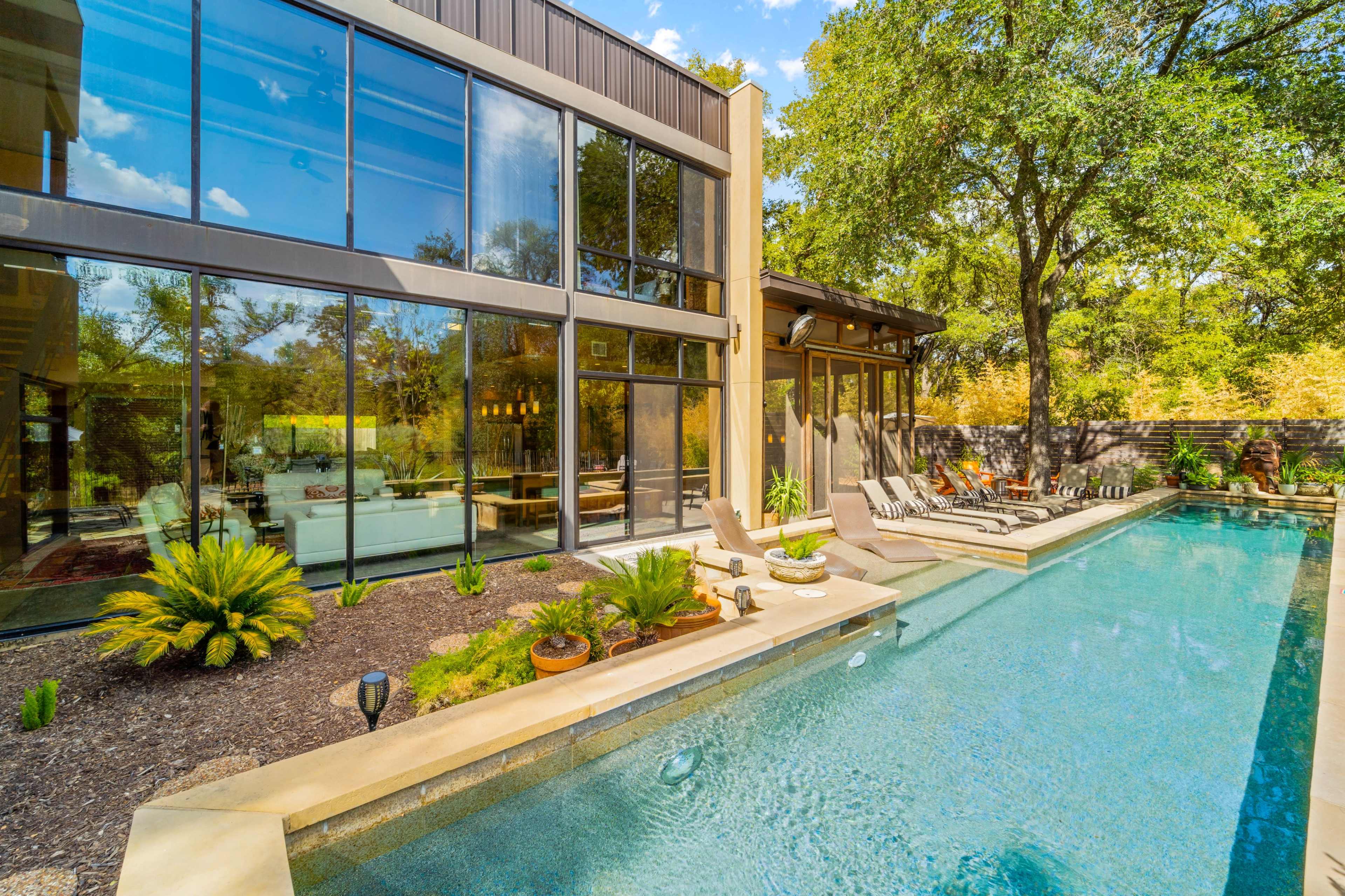 The image shows a modern house with large glass windows overlooking a swimming pool surrounded by landscaping and lounge chairs.