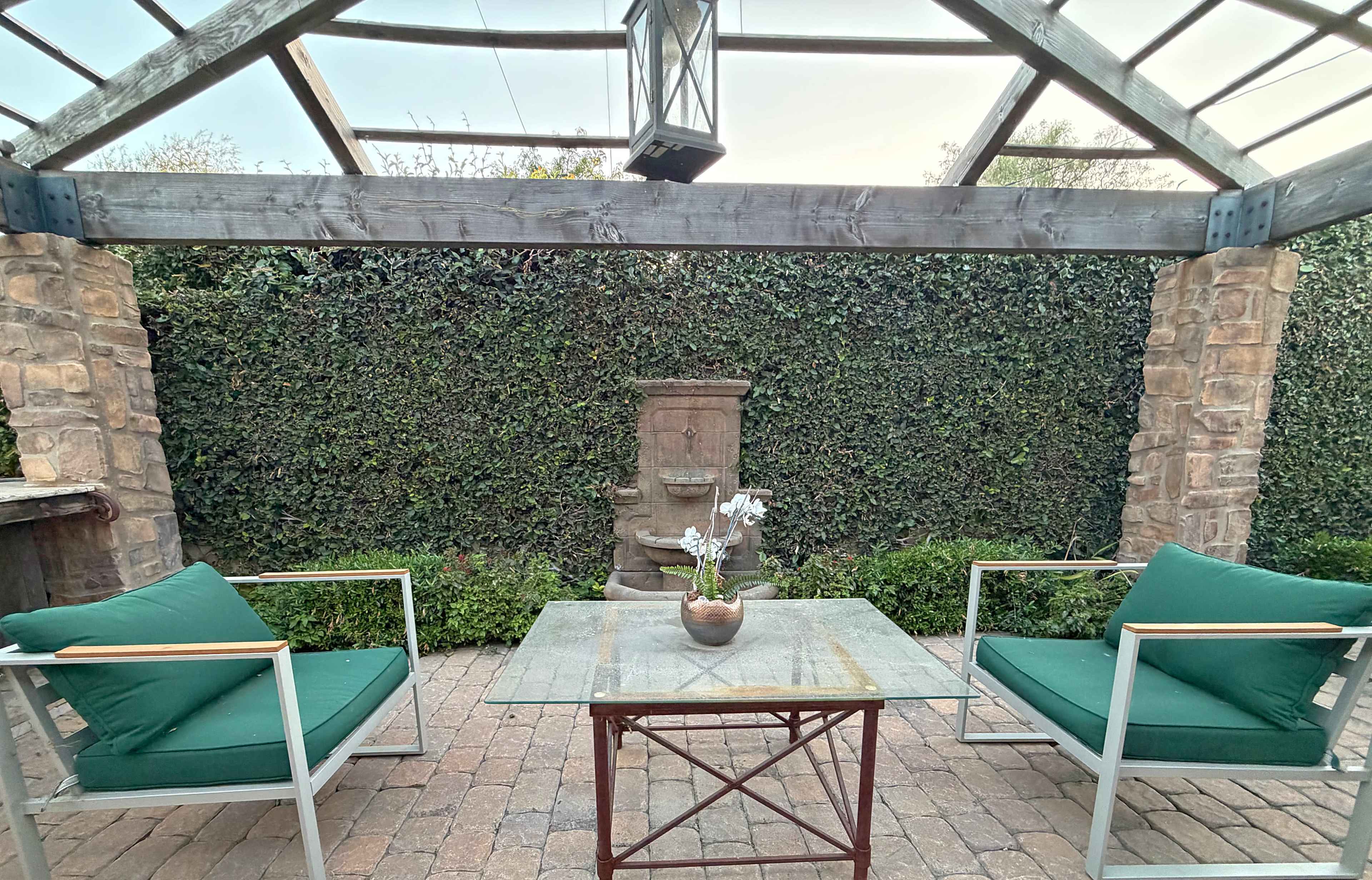 A patio with two green chairs and a glass table is set beneath a wooden pergola, facing a stone wall with a fountain.