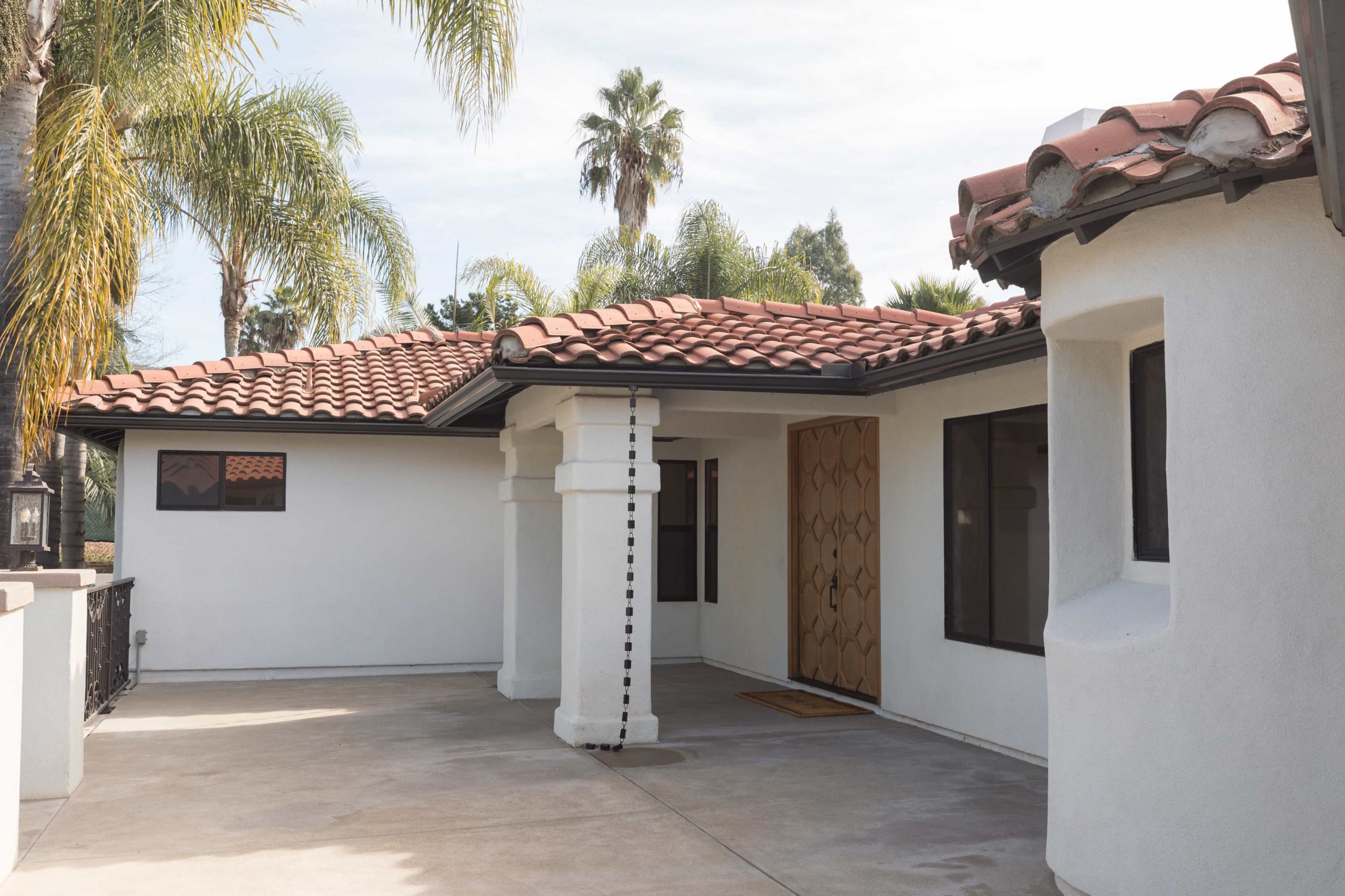 The image shows a single-story home with a tiled roof, a decorative wooden front door, and a spacious patio area surrounded by palm trees.