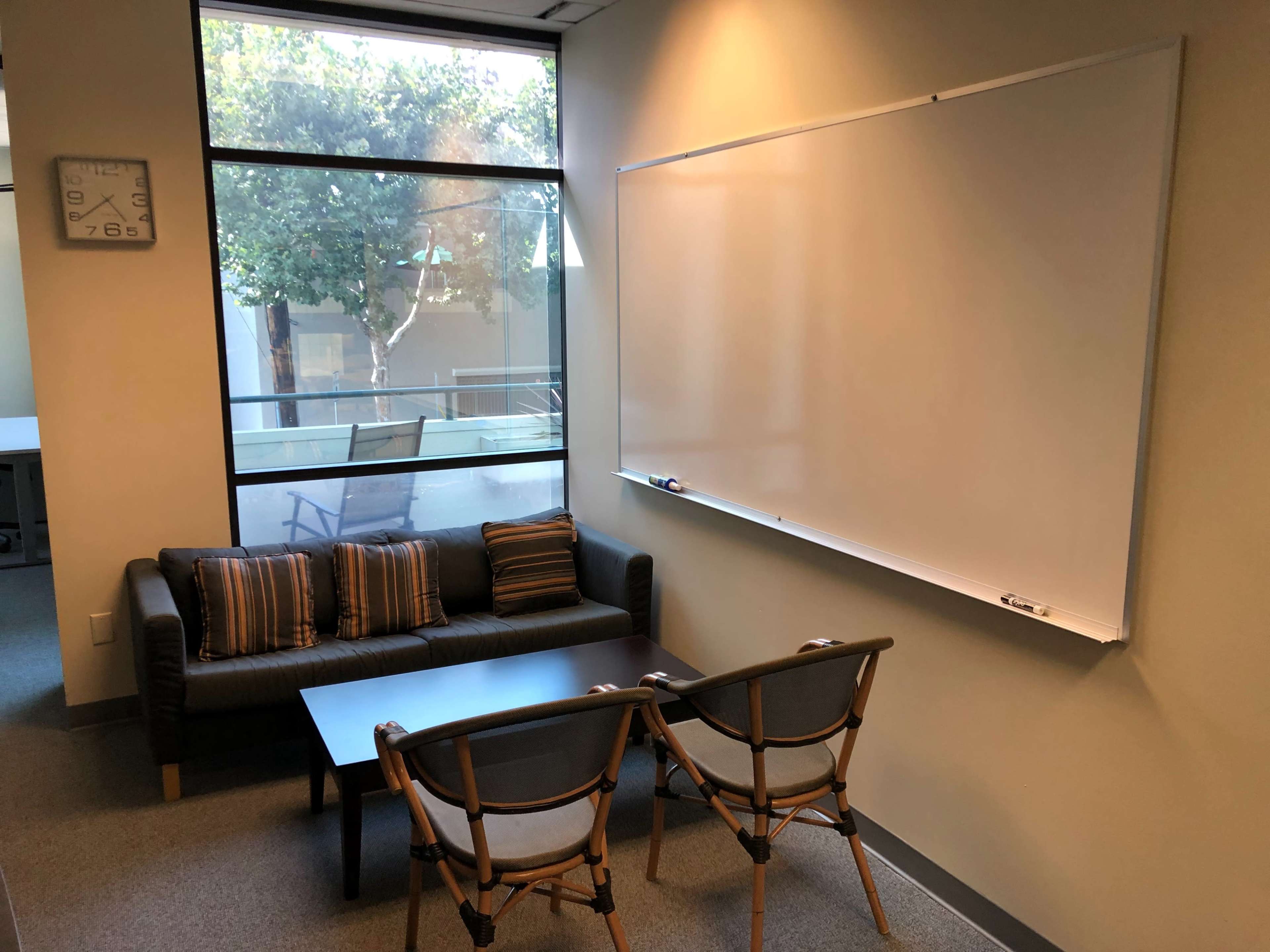 A small meeting room with a couch, two chairs, a coffee table, a whiteboard, and a large window letting in natural light.