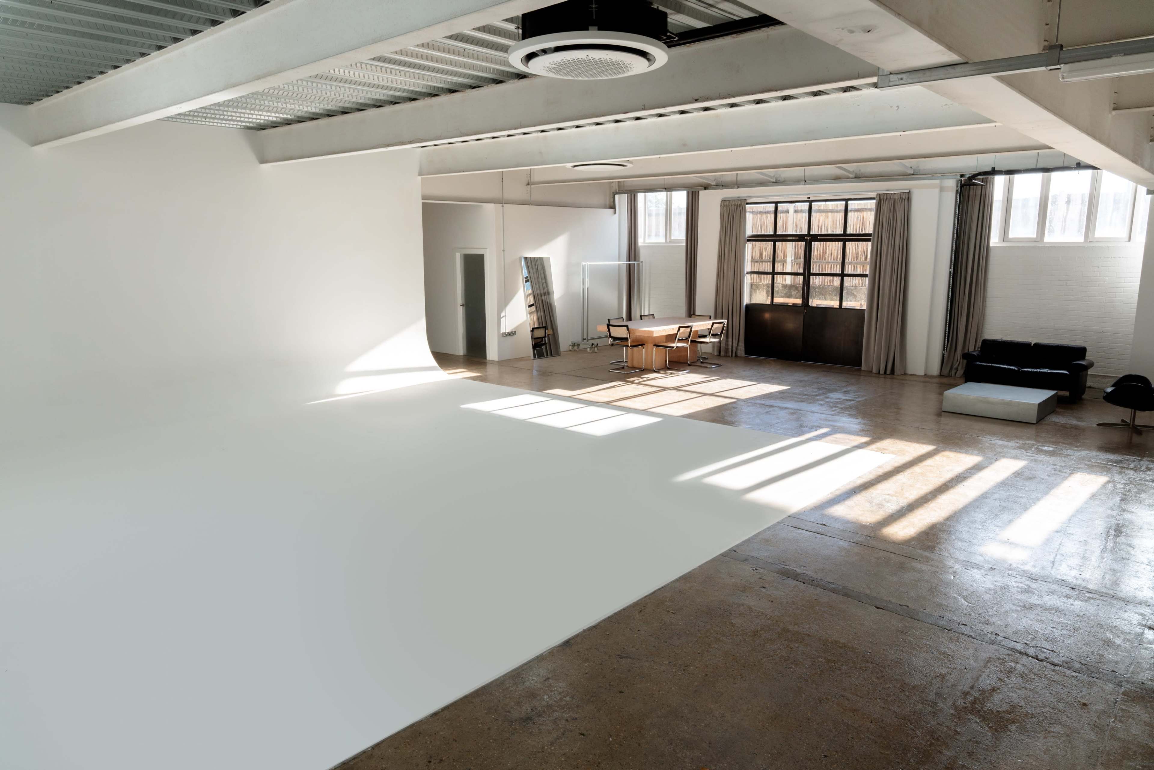 The image shows a spacious, well-lit studio with a white floor, large windows, a table with chairs, and a sofa against one wall.