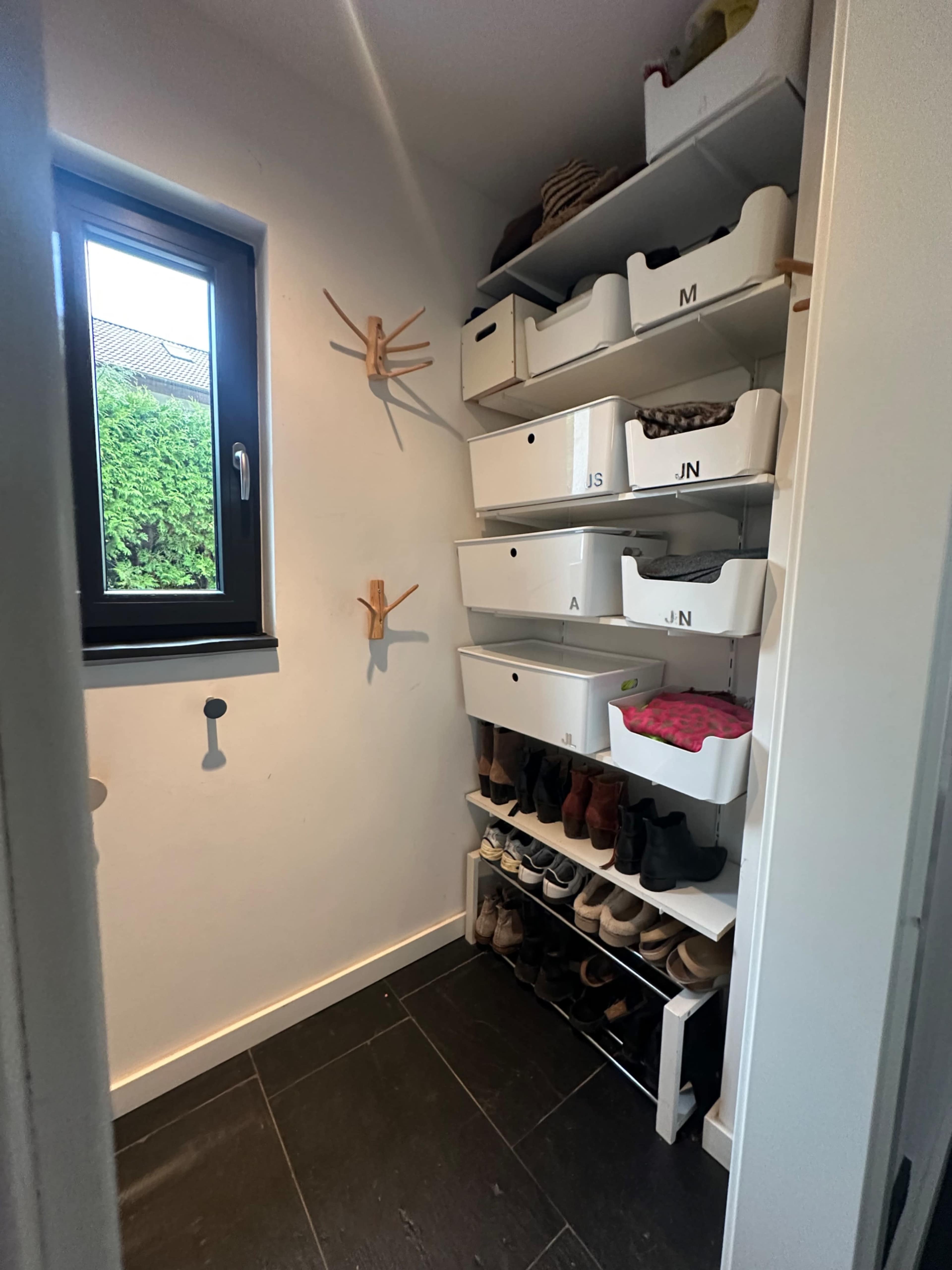 The image shows a small storage corner with a window, featuring shelves with labeled white boxes and a row of shoes at the bottom.