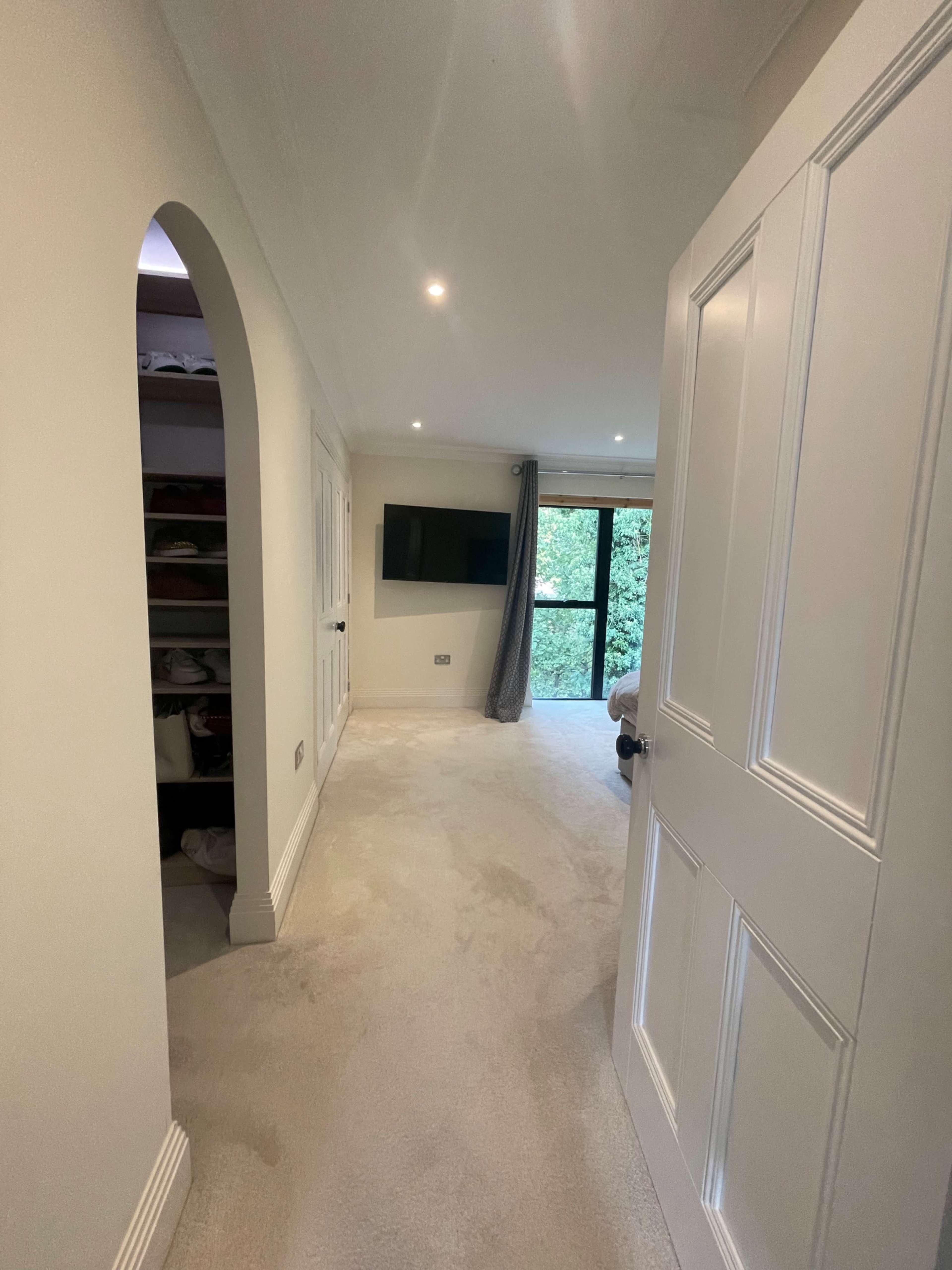 The image shows a well-lit hallway leading to a room with a television mounted on the wall and a view of greenery outside through a window.
