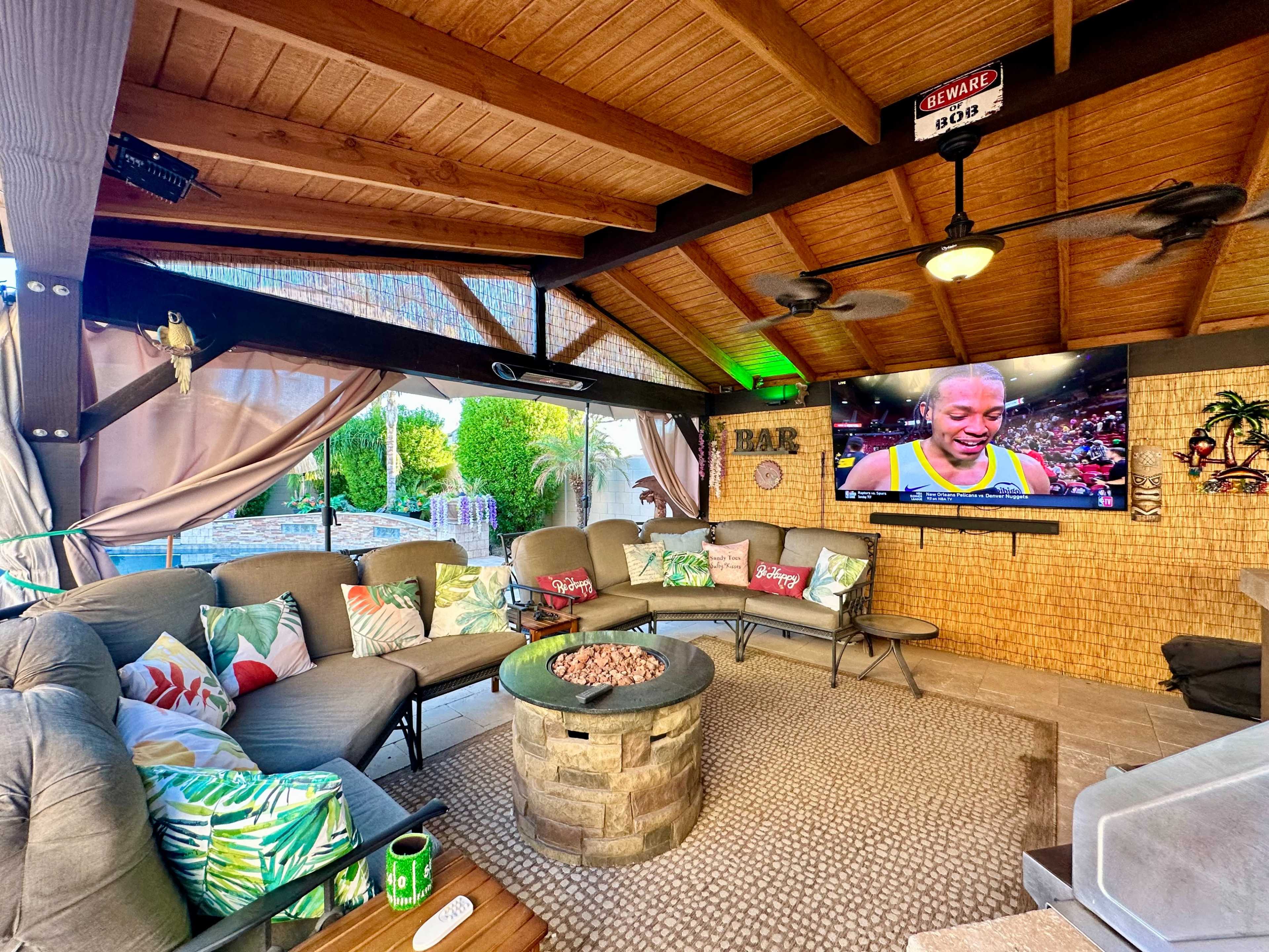A covered outdoor seating area with a circular stone fire pit, a large television displaying a sports event, and comfortable furniture featuring decorative cushions.