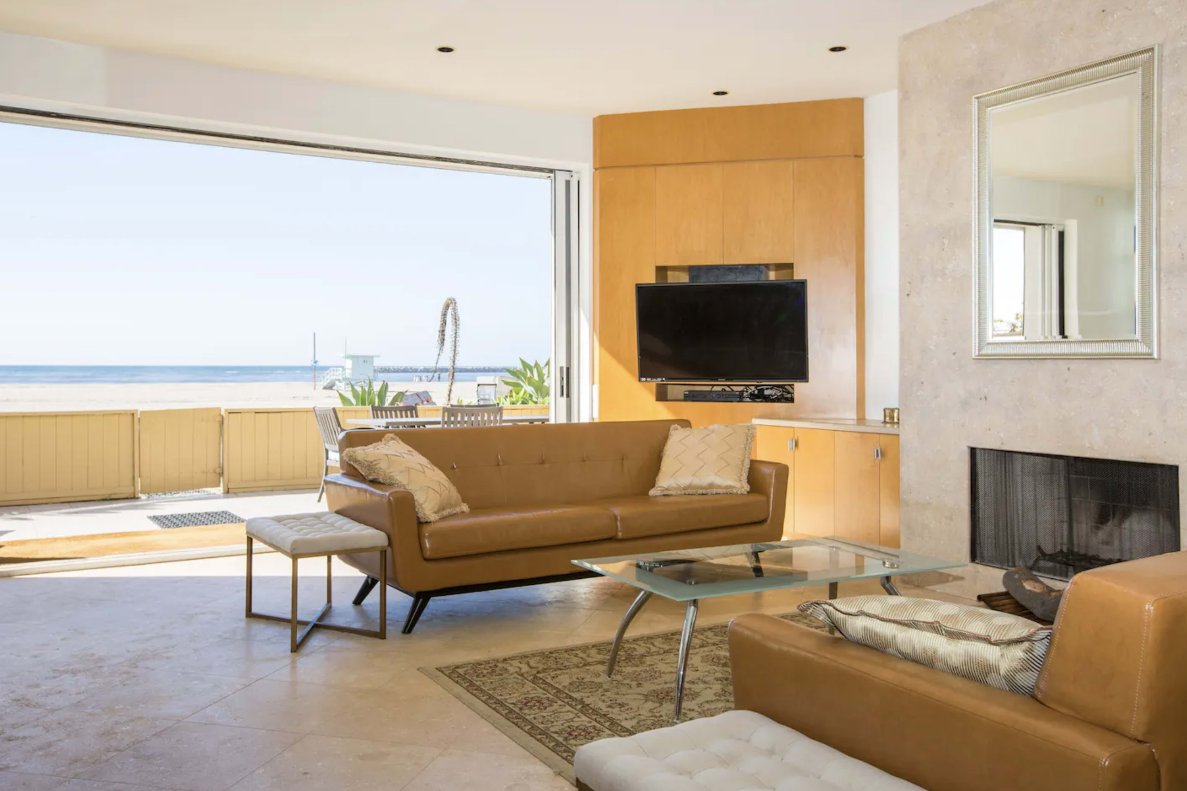 A modern living room features a brown leather sofa, a glass coffee table, and large sliding doors opening to a view of the ocean.
