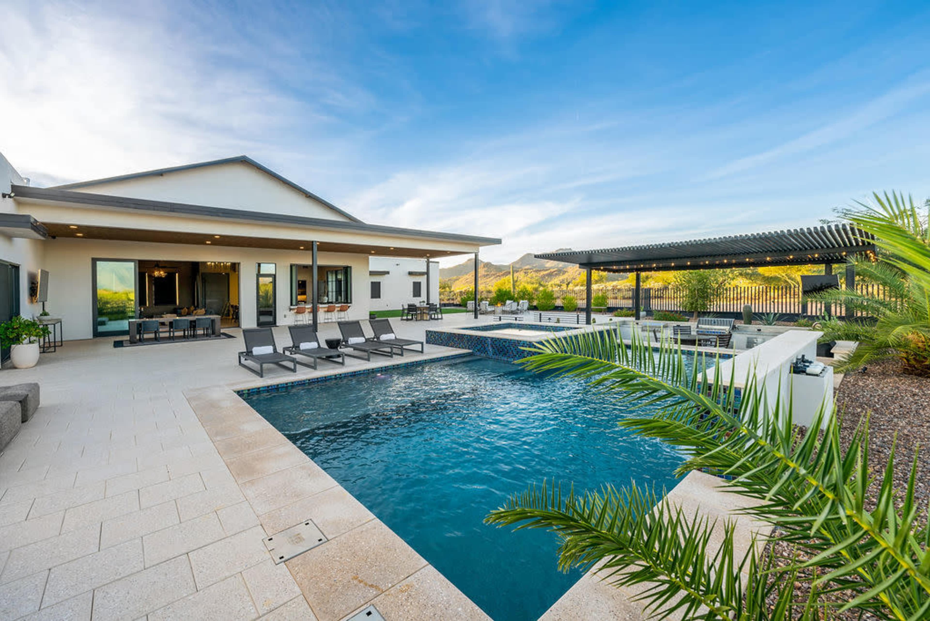 The image shows a modern outdoor pool area with lounge chairs, a covered seating space, and landscaped surroundings against a backdrop of mountains.