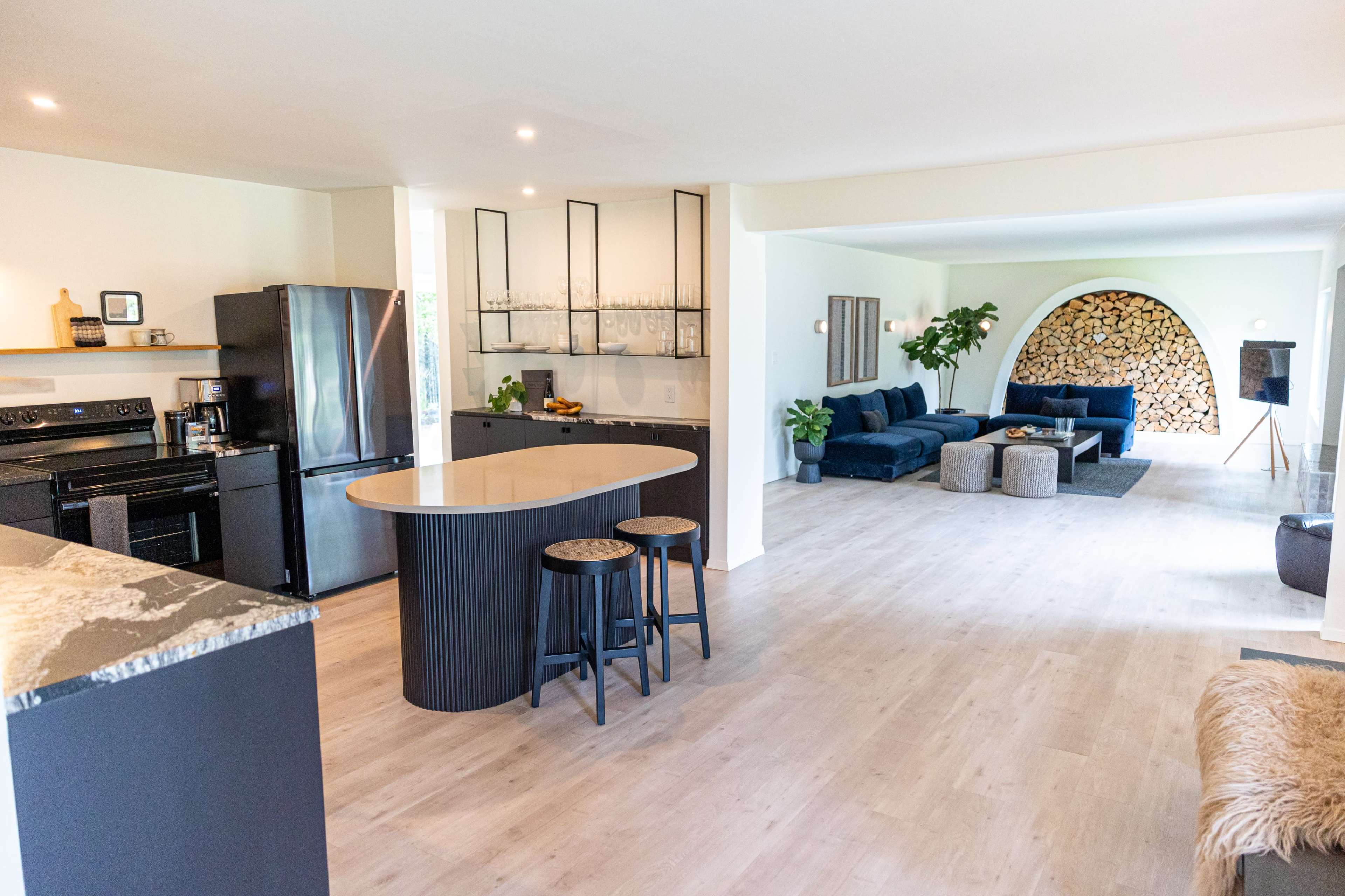A modern, open-concept kitchen and living area featuring sleek black appliances, a central island with bar stools, and a large sectional sofa placed near a stacked log feature.