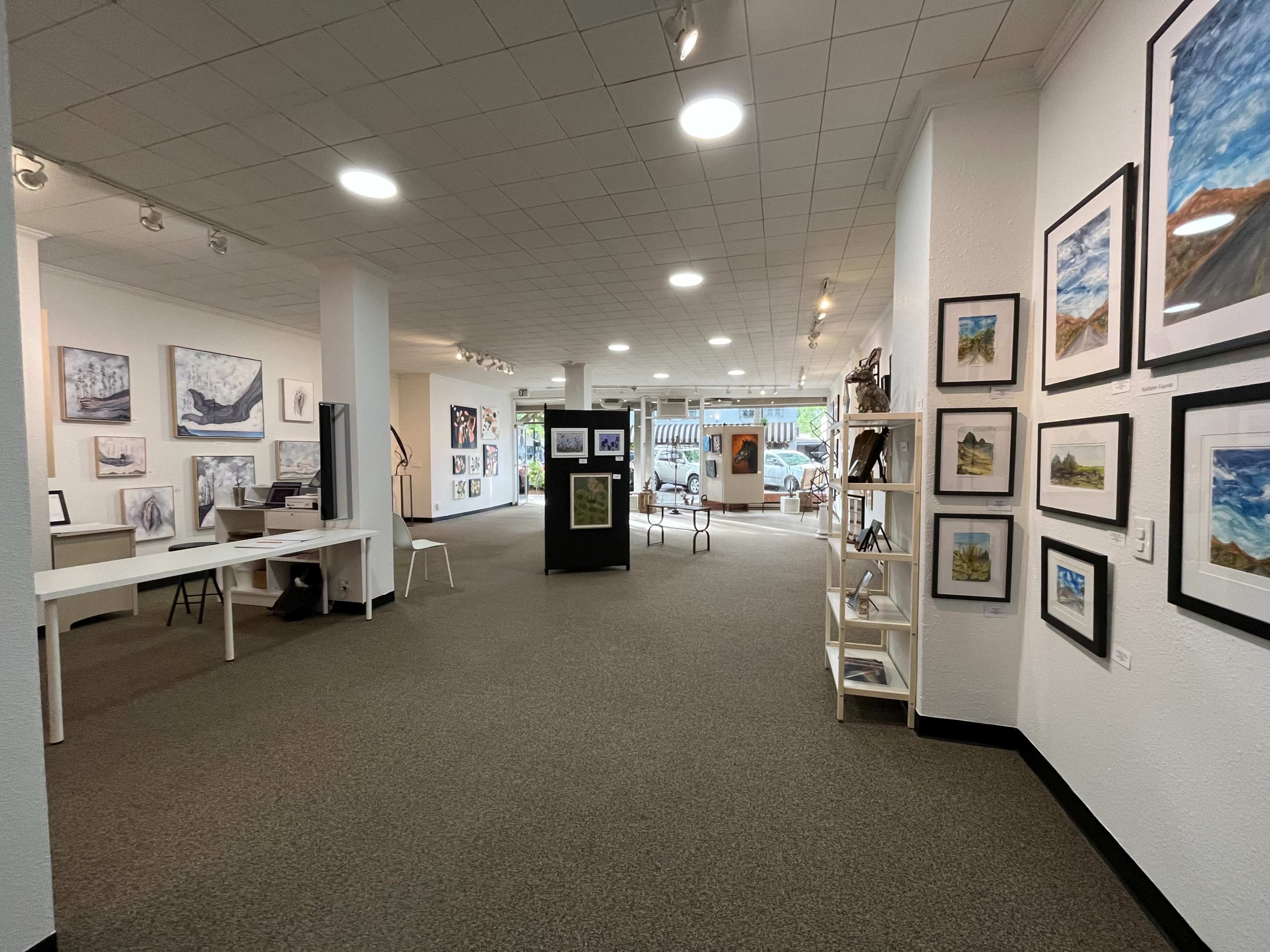 The image shows a spacious art gallery with white walls displaying various framed artworks, while tables and seating areas are arranged throughout the room.