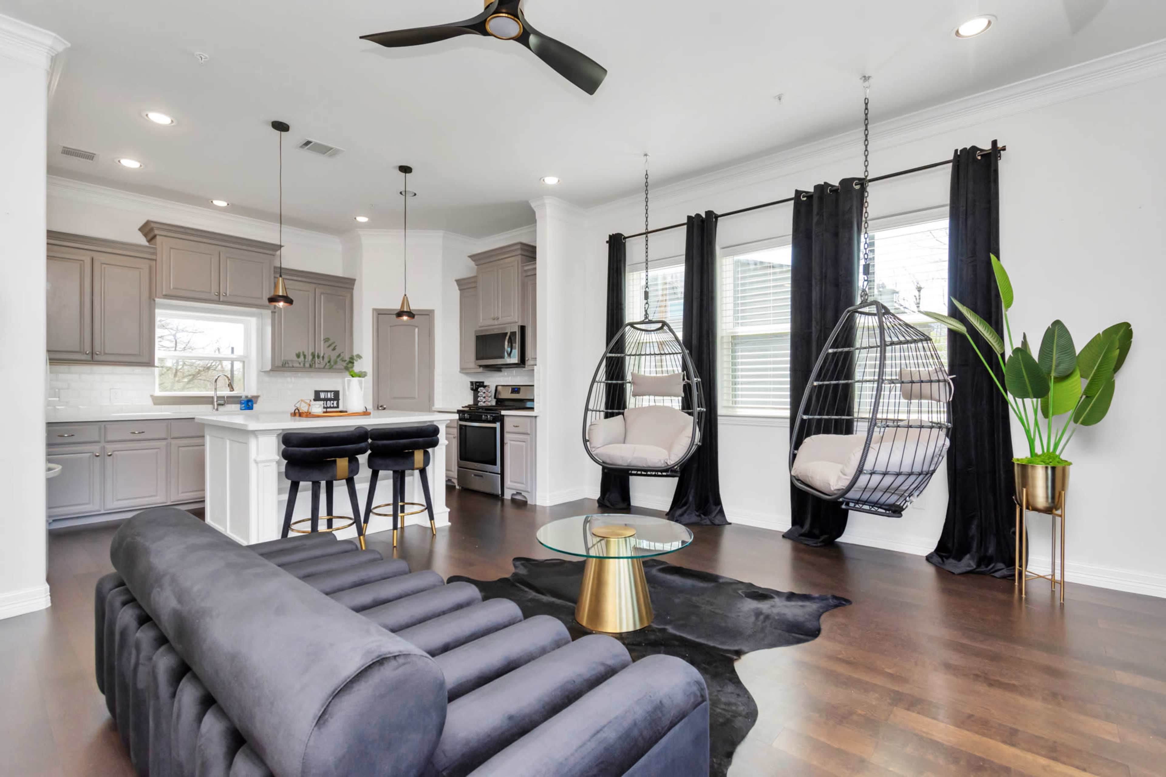 A modern living area with a gray sofa, two hanging chairs, a glass coffee table, and a kitchen in the background.