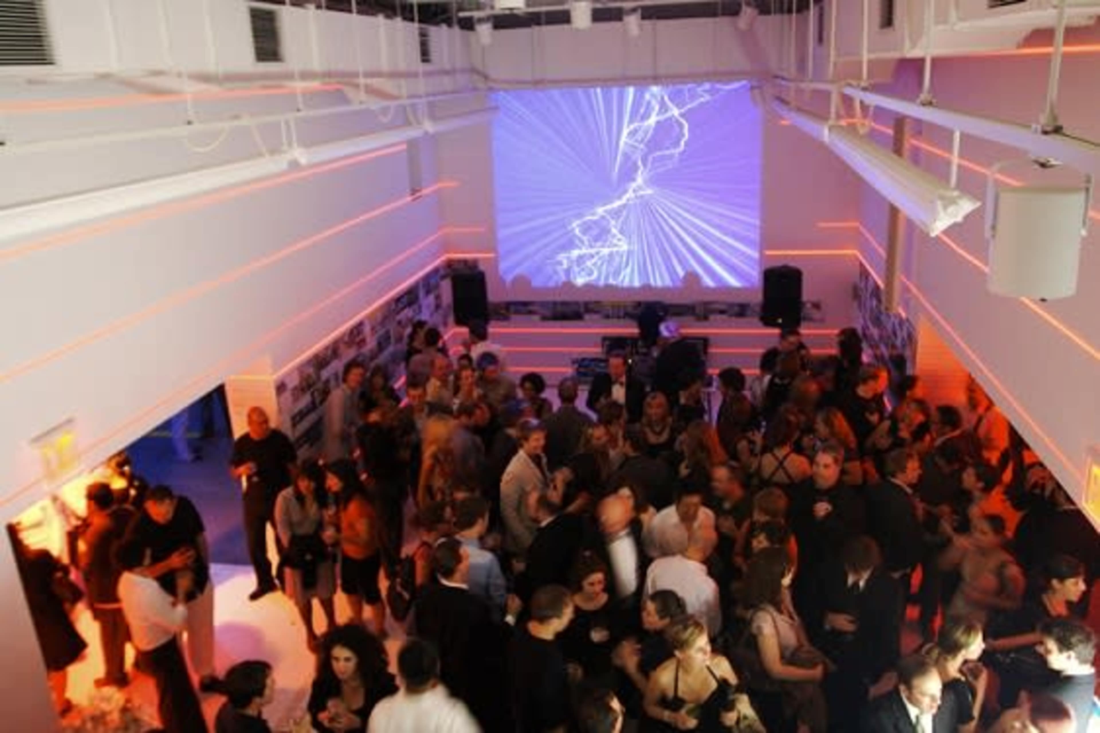 A large crowd mingles at a modern indoor event space featuring a DJ booth and a colorful light display on the wall.