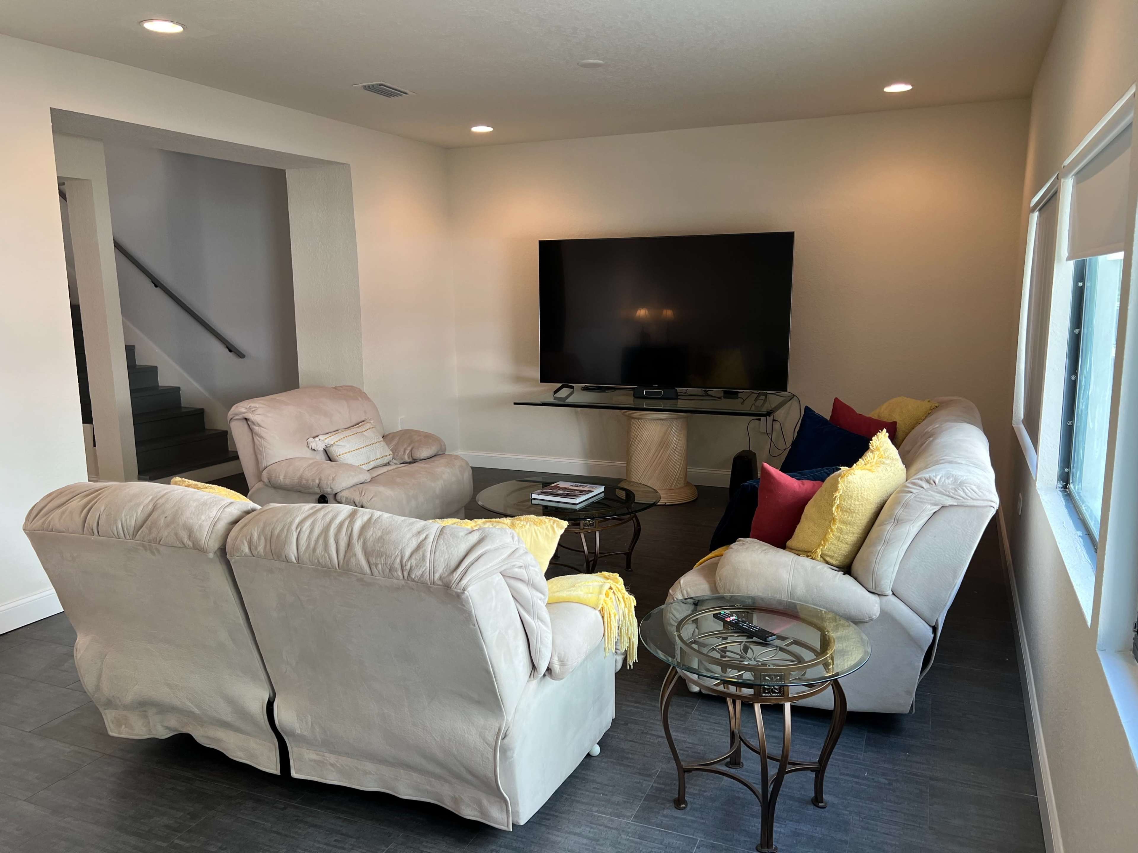 A modern living room features light-colored sofas, a glass coffee table, and a large television mounted on the wall.