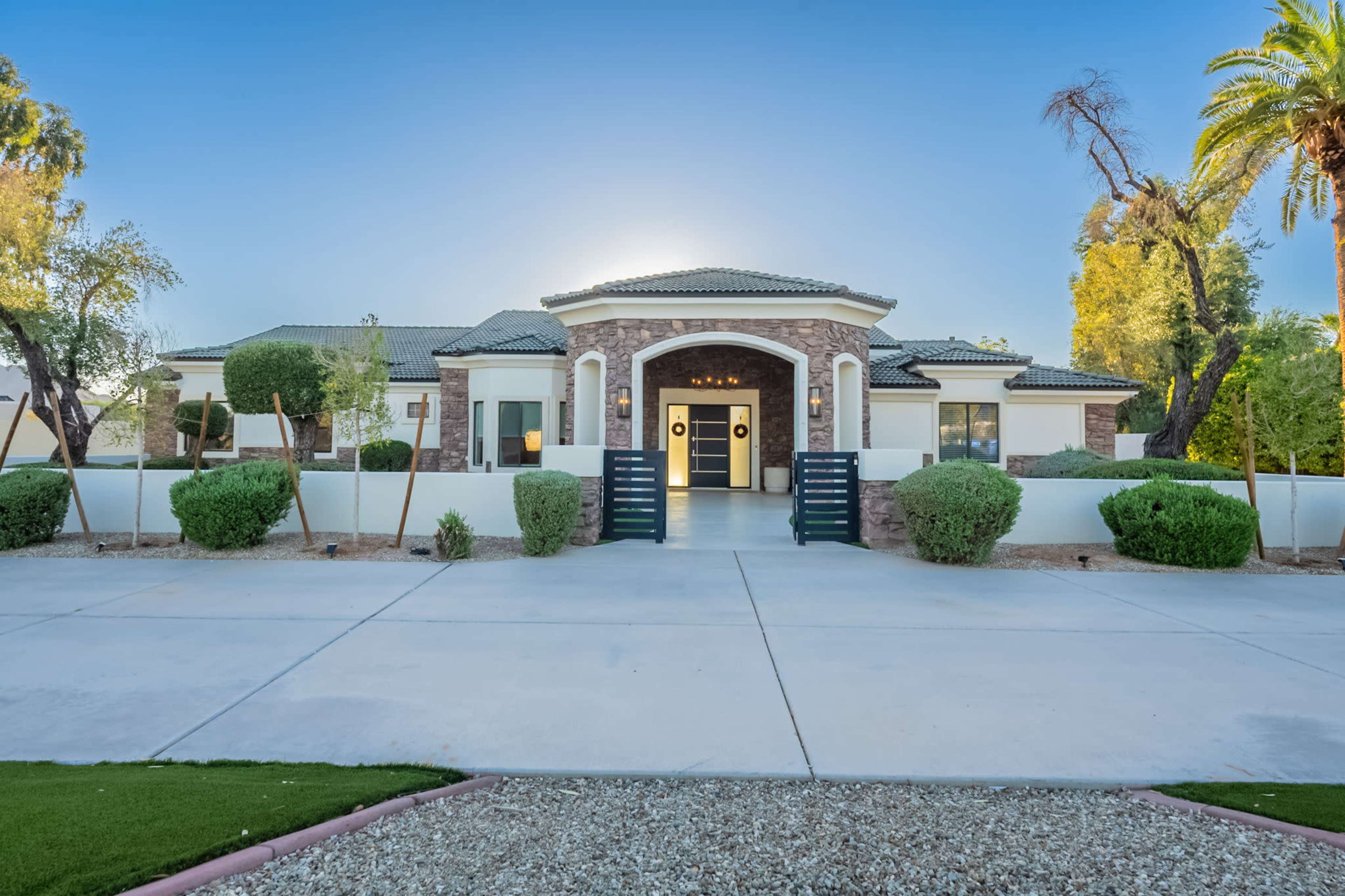 The image shows a modern single-story house with a circular driveway, surrounded by landscaped greenery and palm trees.