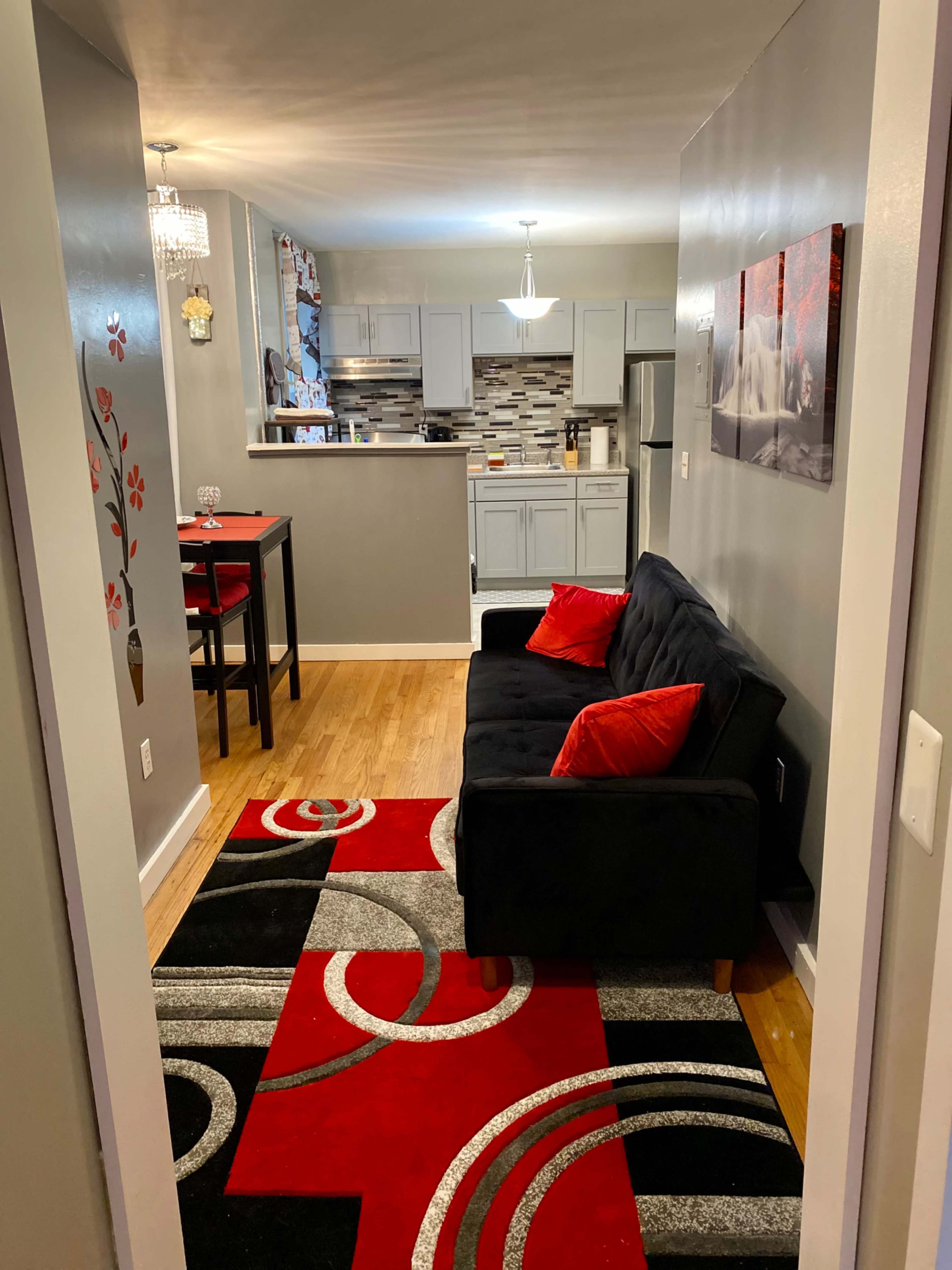 The image shows a small living area featuring a black sofa with red cushions, a patterned rug, and a view of a kitchen area with modern cabinetry and a dining table.