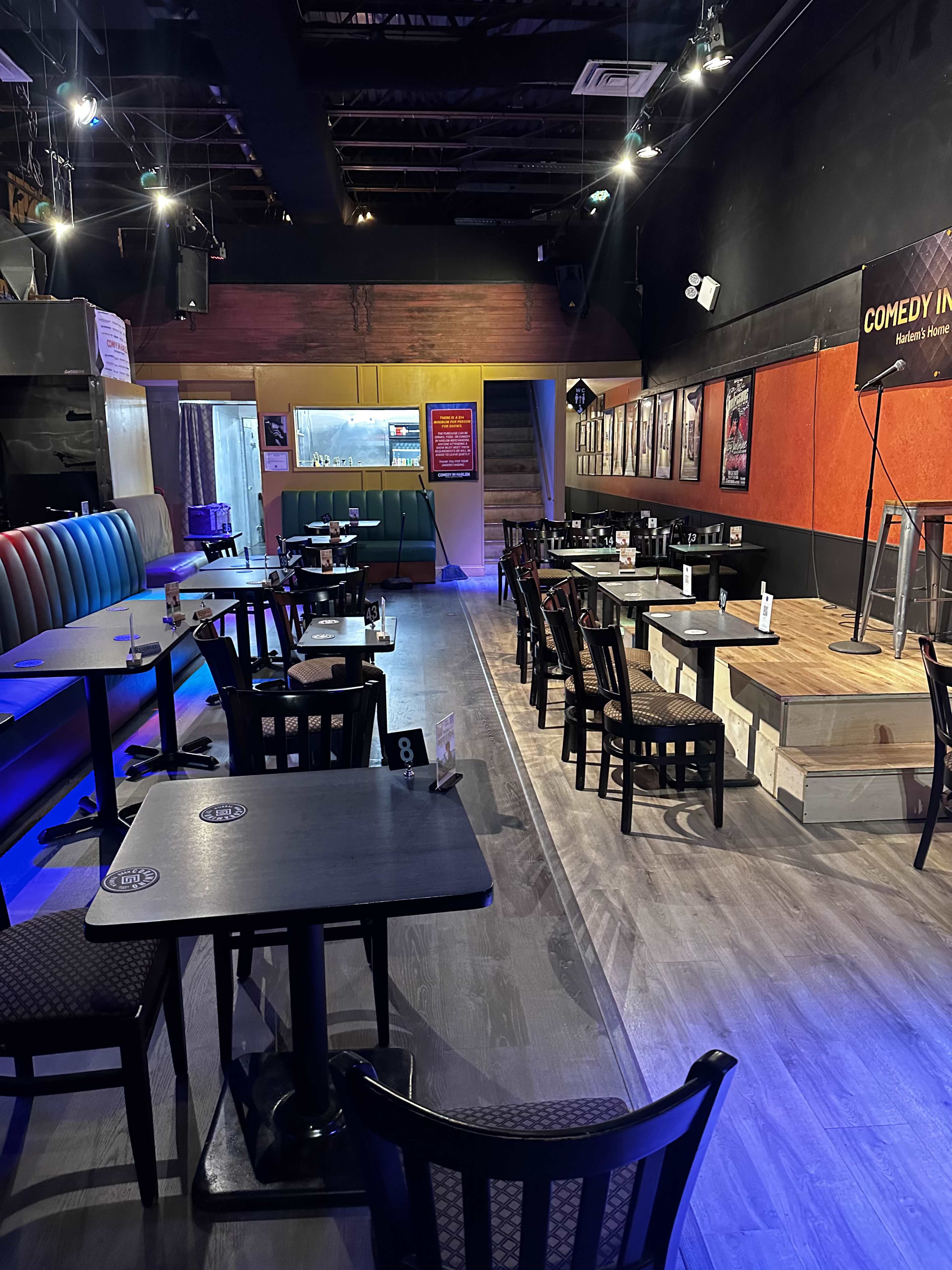 The image shows a dimly lit comedy club with empty tables and a small stage at the front.