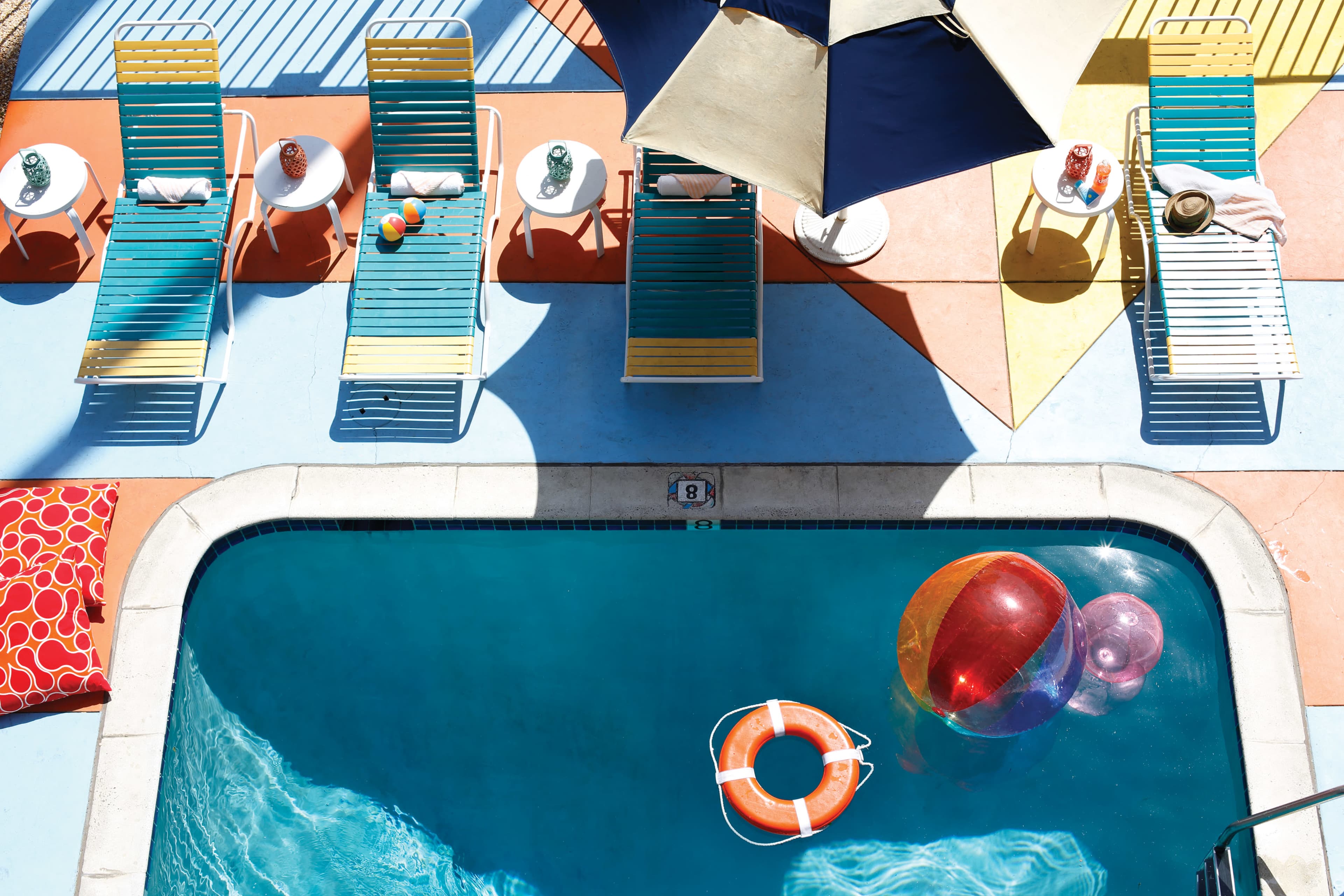 A swimming pool surrounded by colorful lounge chairs, tables, and pool floats under a large umbrella.