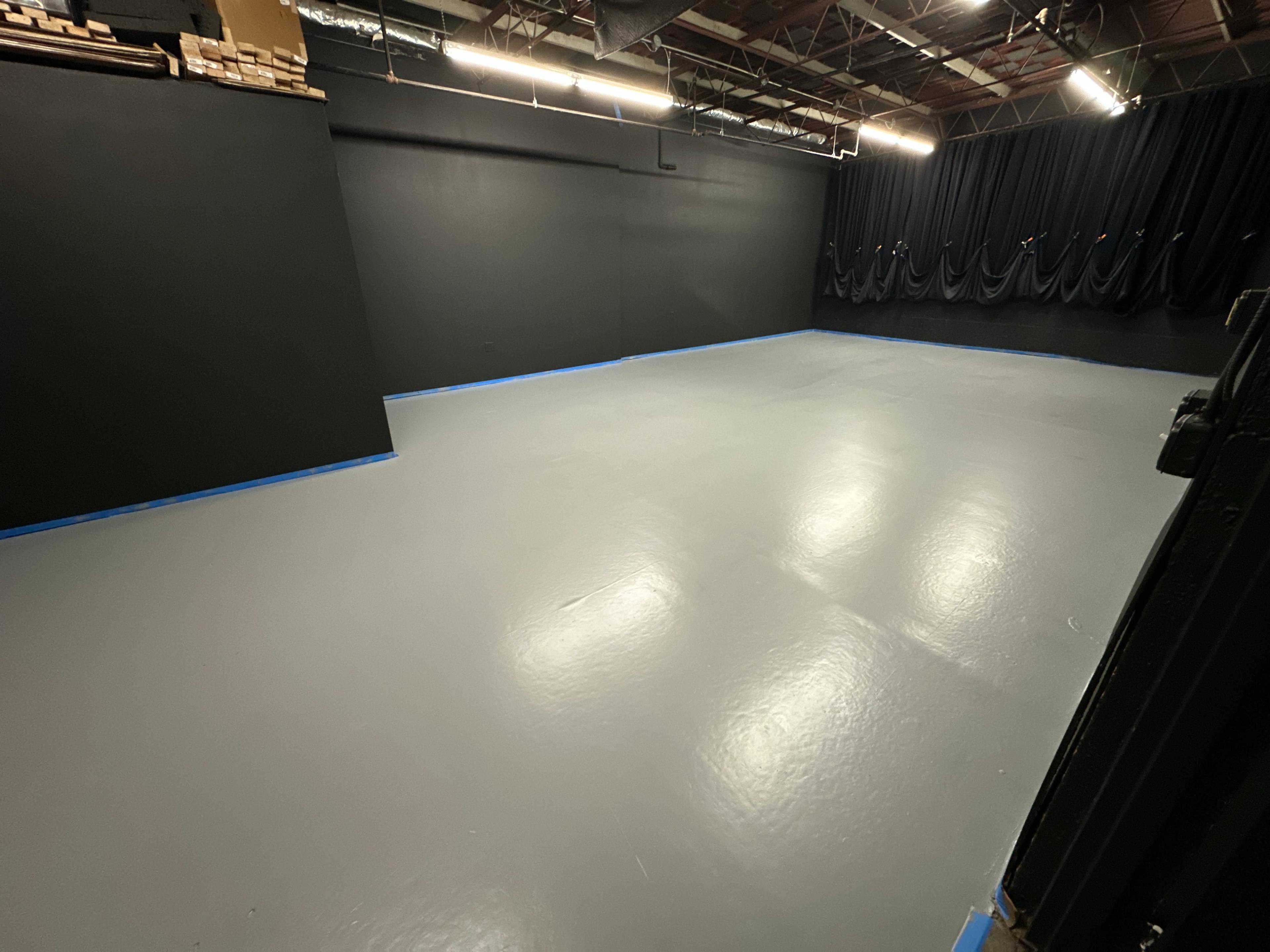 The image shows a spacious room with a gray polished floor and black walls, featuring blue tape along the edges.