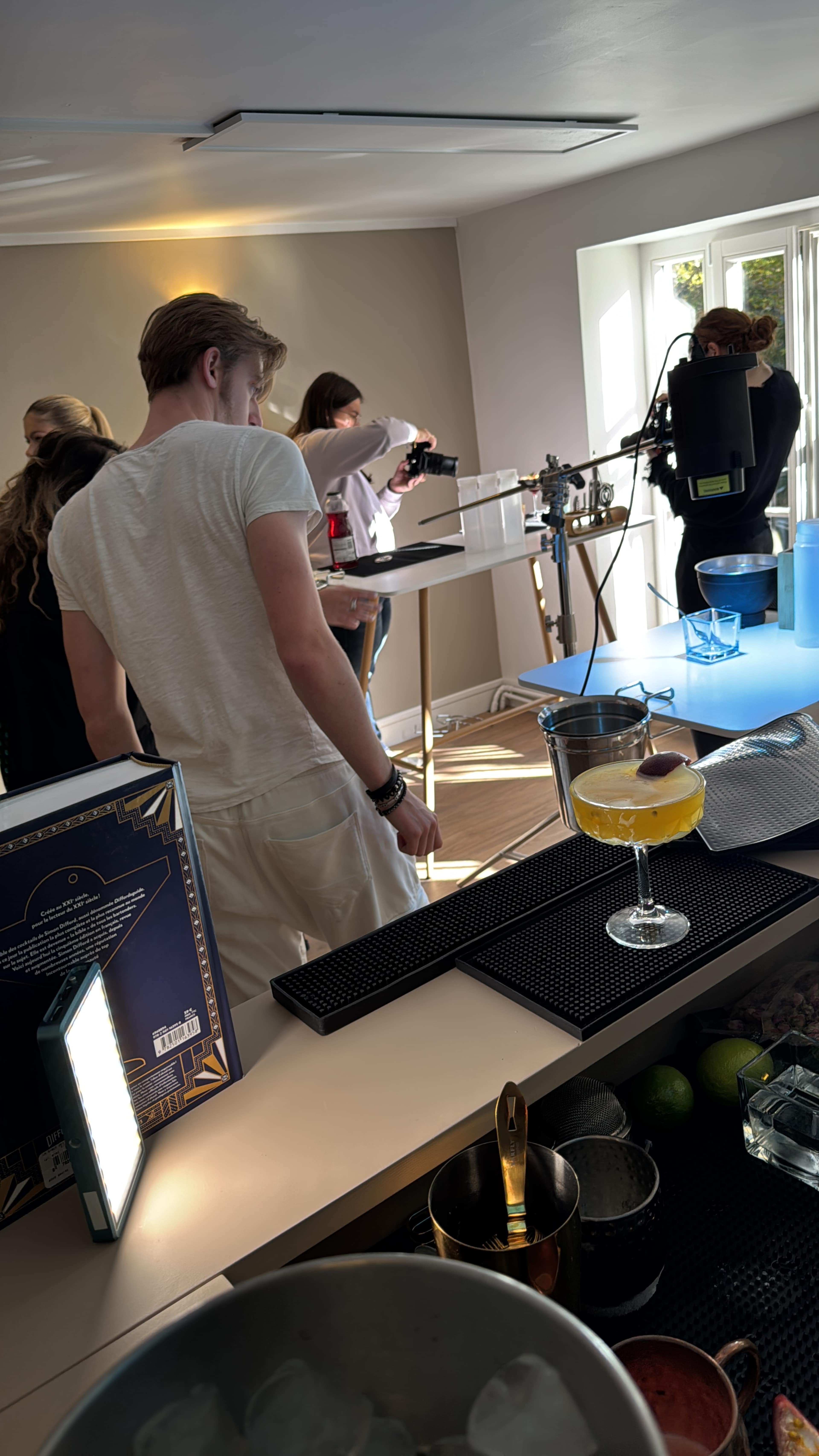 A group of people is preparing cocktails at a well-lit bar setup, with cameras and equipment positioned for filming.