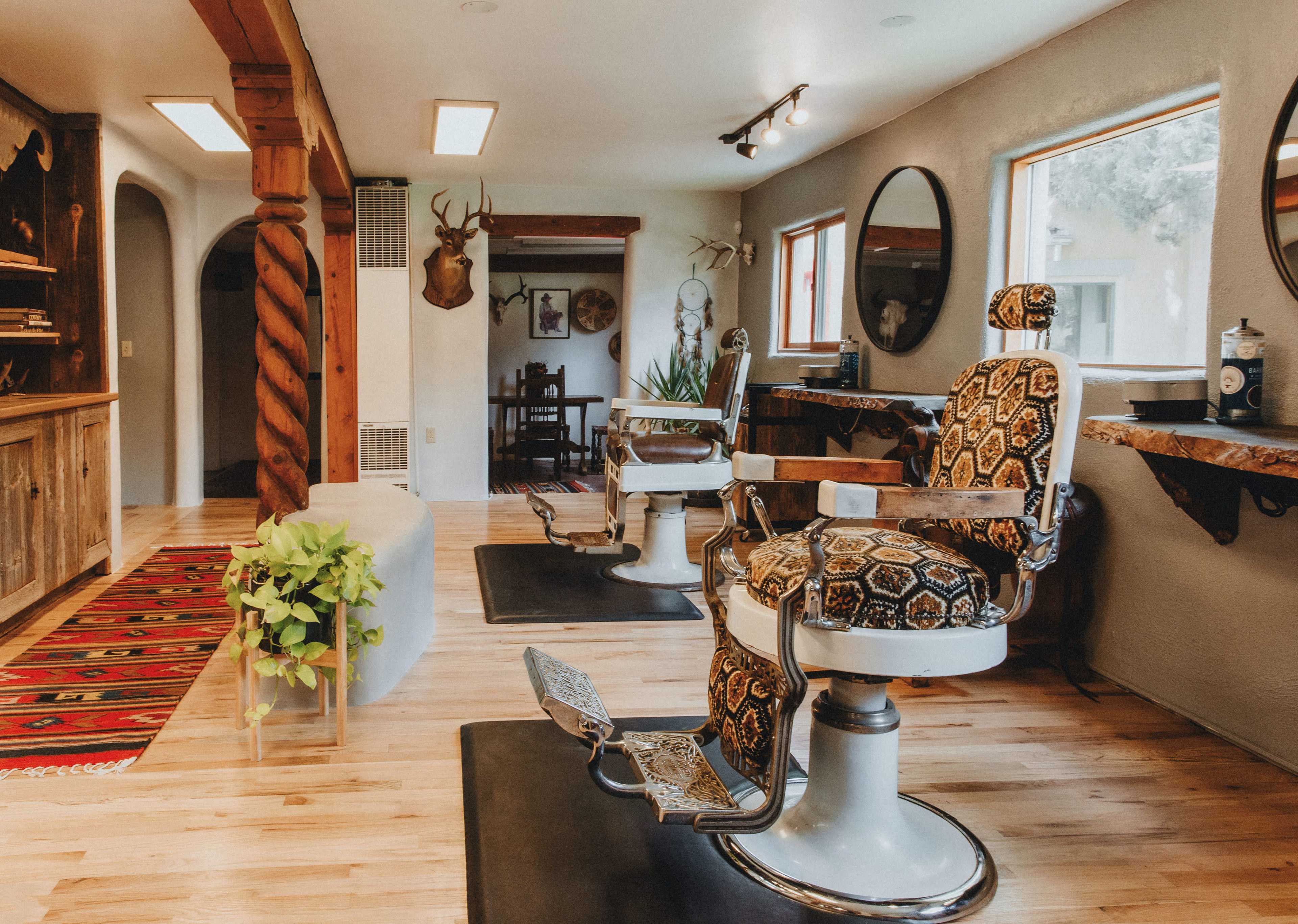 The image shows a stylish barbershop interior featuring two vintage barber chairs with patterned upholstery, wooden beams, and natural light streaming through large windows.