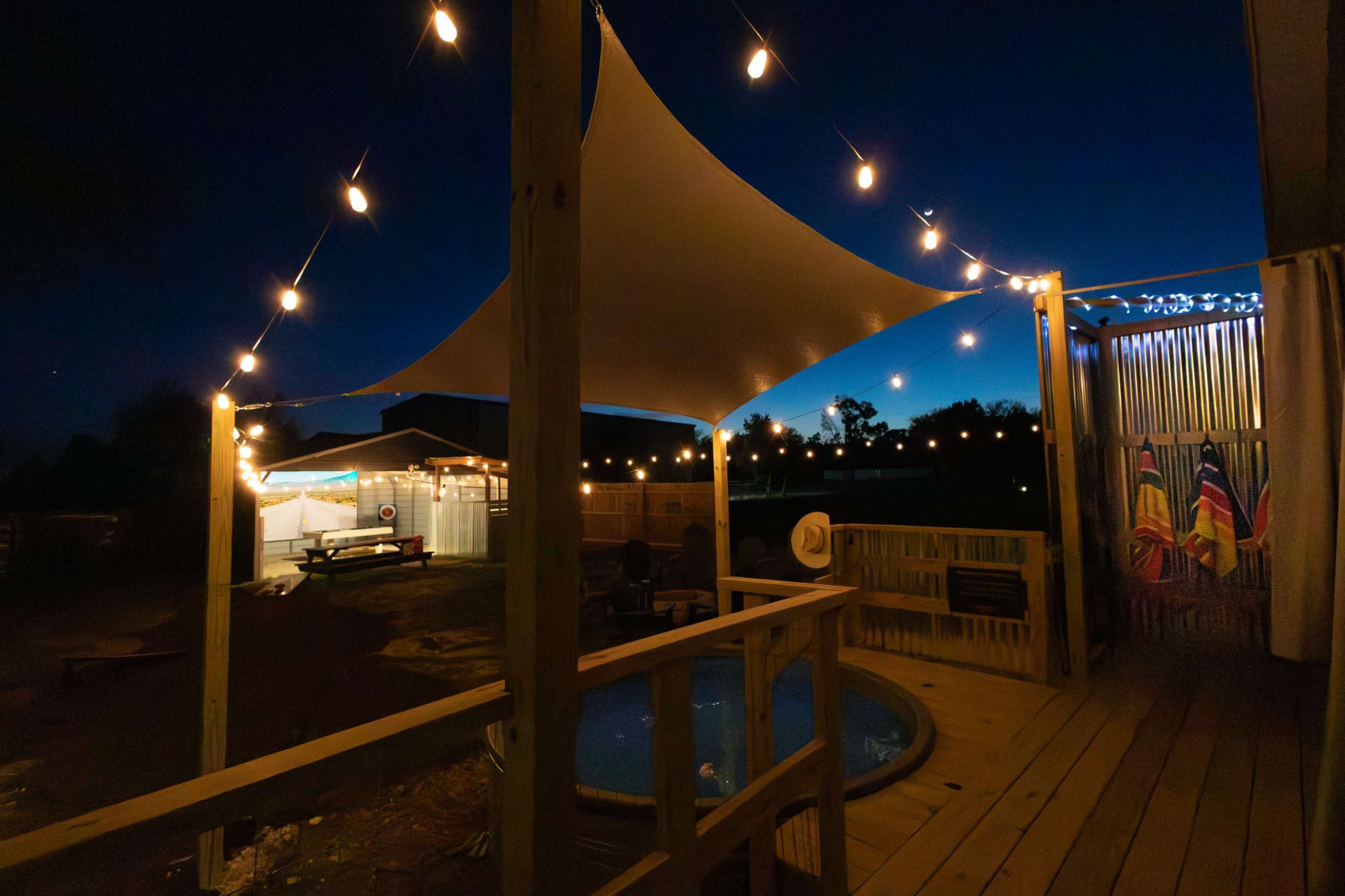 A deck area with a pool features a large sail canopy and string lights, with a view of a brightly lit outdoor setup in the background at dusk.