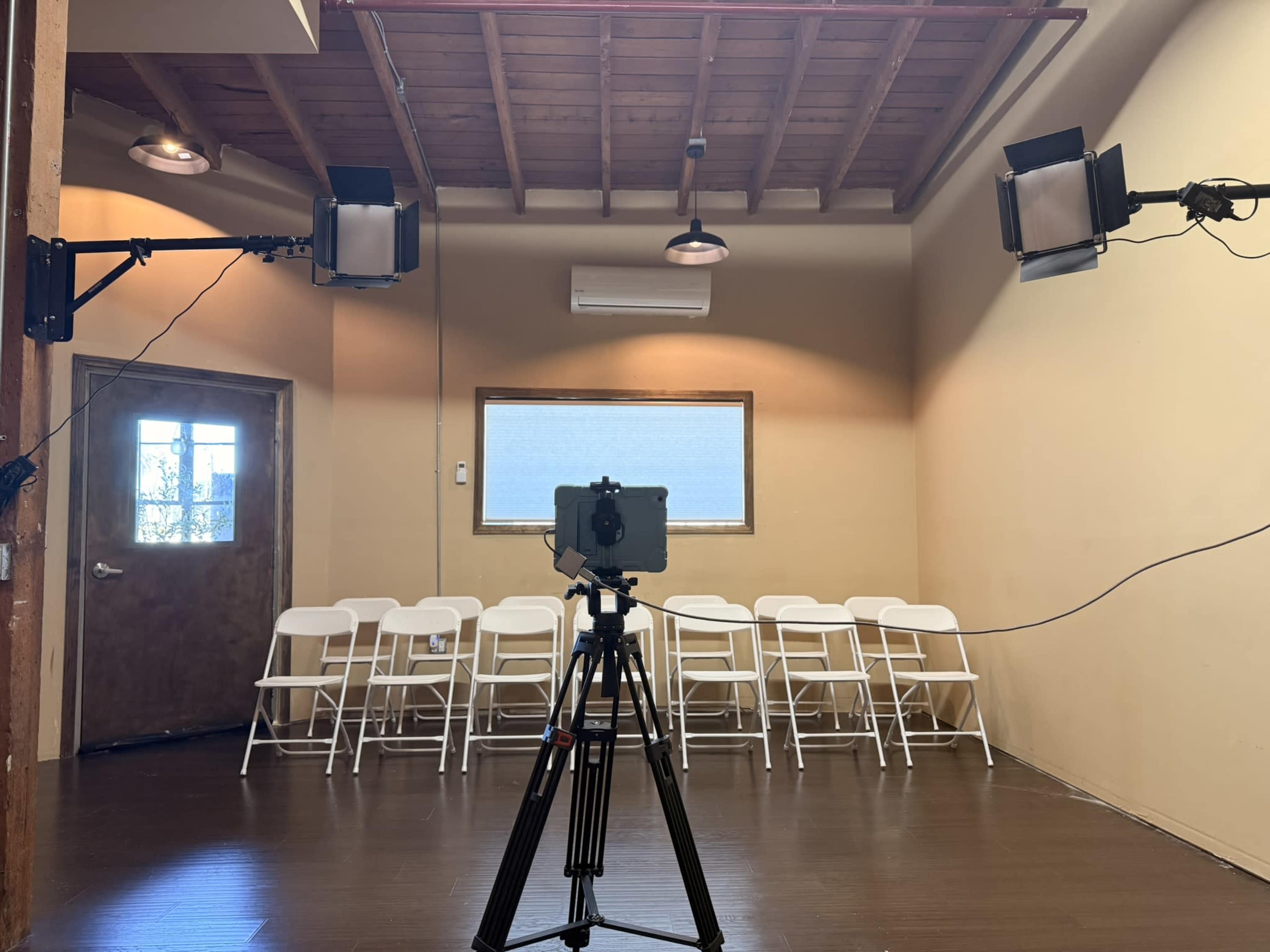 A camera is set up on a tripod facing a row of white chairs in a well-lit room with a wooden ceiling and a door.