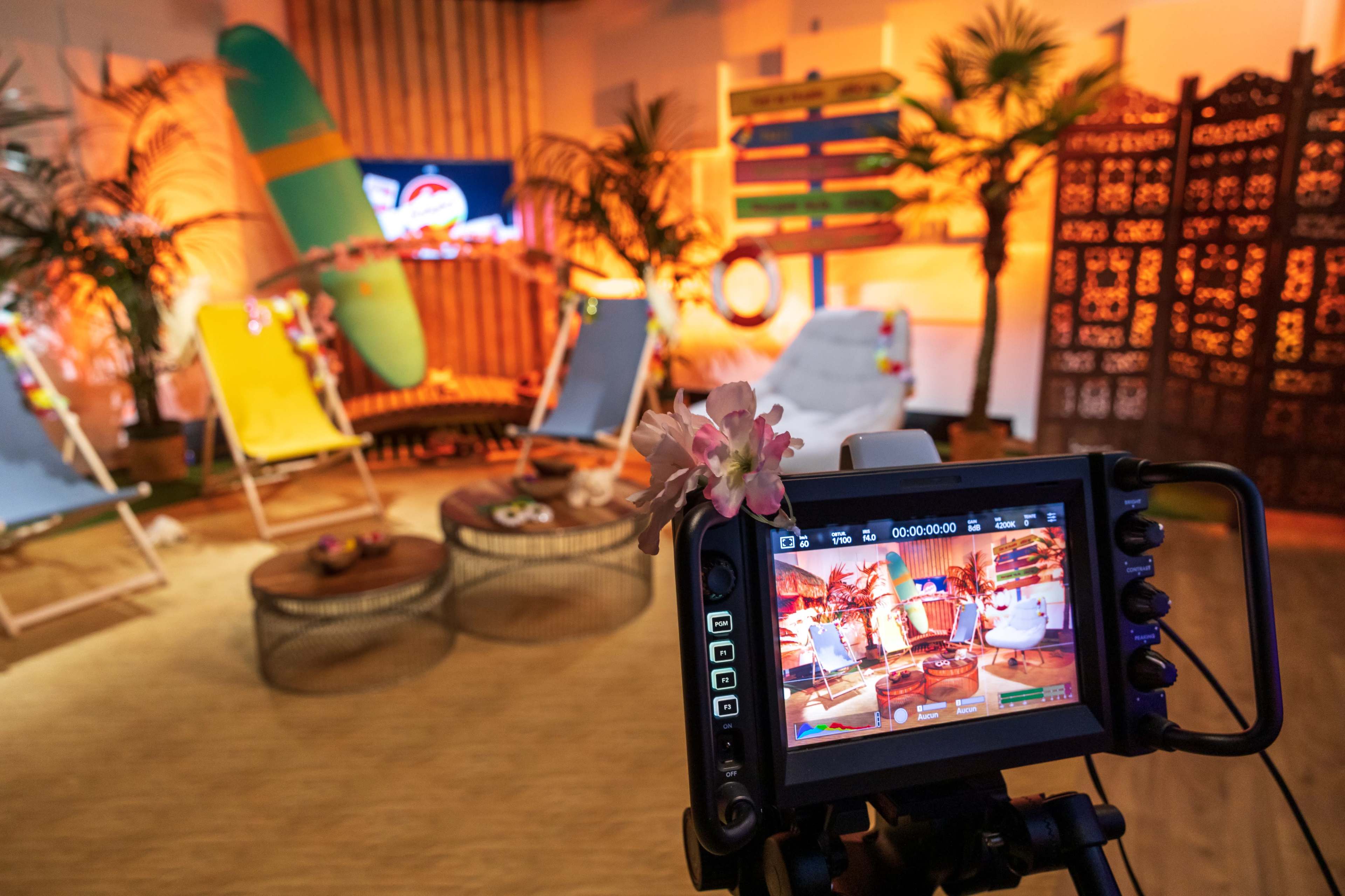 A camera is focused on a brightly lit tropical-themed studio set featuring beach chairs, decorative plants, and a surfboard.