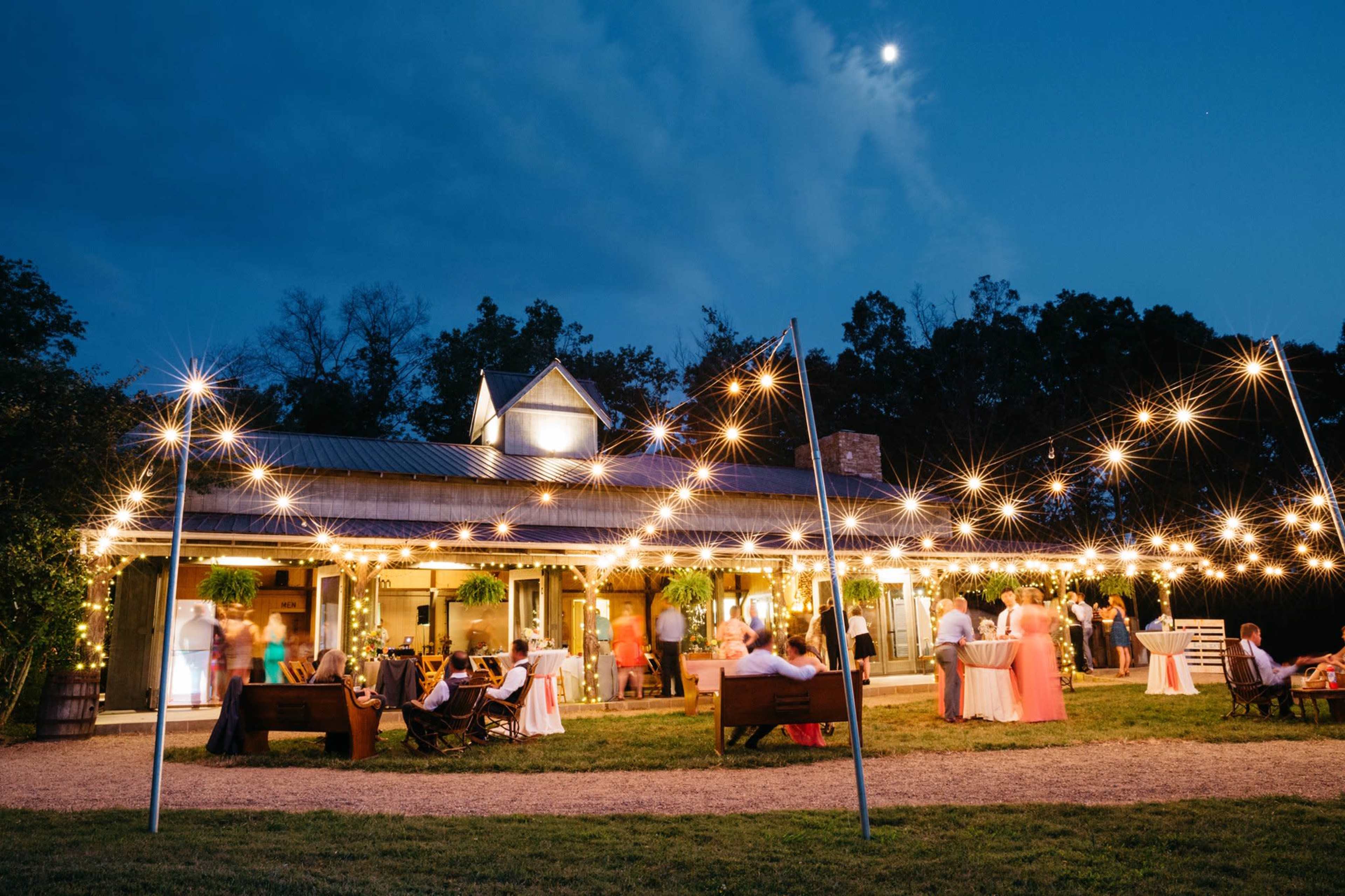 A gathering is taking place outdoors at a venue illuminated by string lights, with guests mingling near tables and a clear sky visible at dusk.