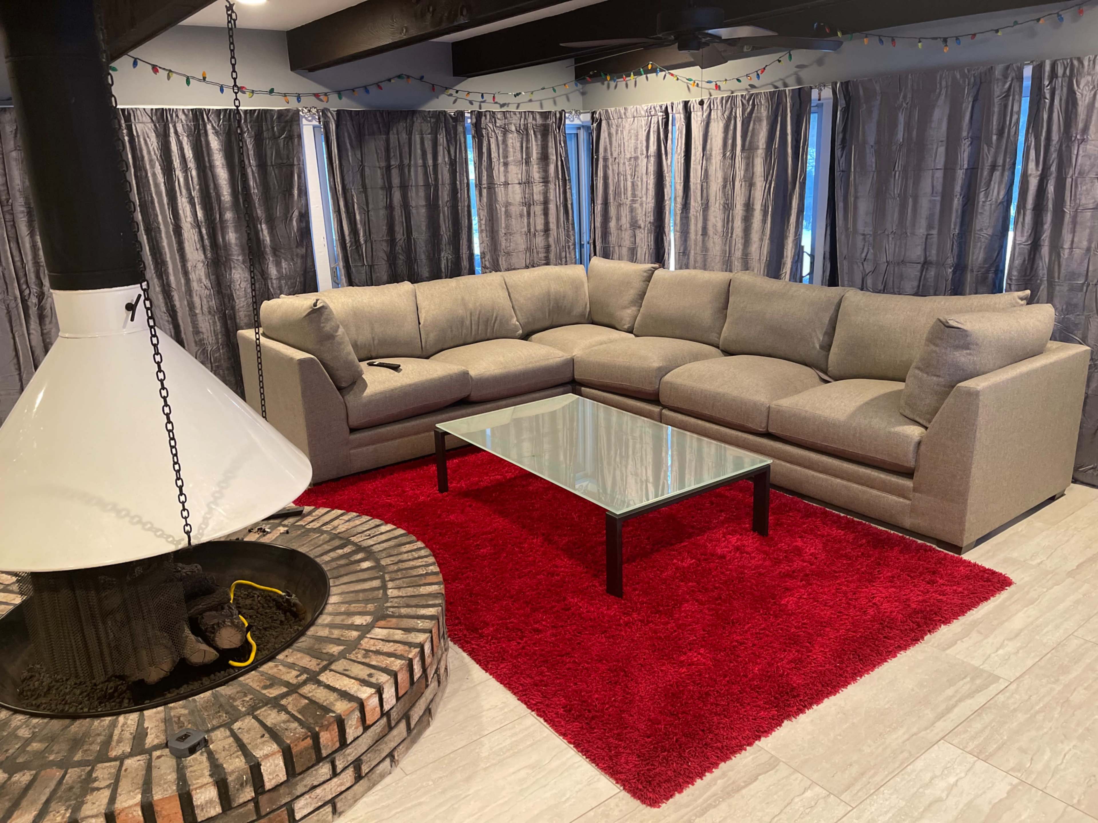 A living room with a large gray sectional sofa, a glass coffee table, and a circular fireplace surrounded by brick, all set on a red area rug.