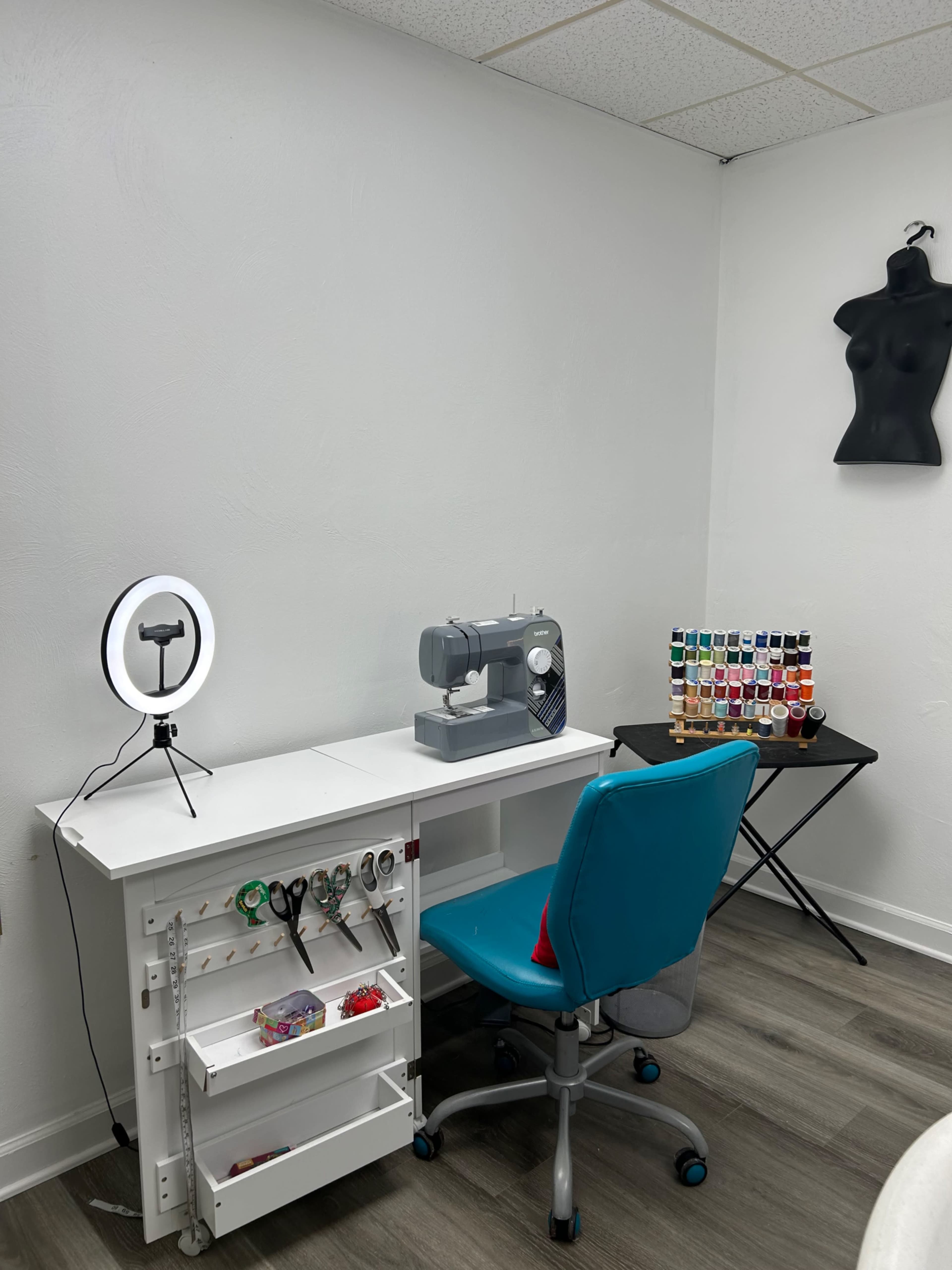 A sewing workspace featuring a sewing machine on a white desk, a blue office chair, a ring light, and a color assortment of threads arranged on a black folding table.