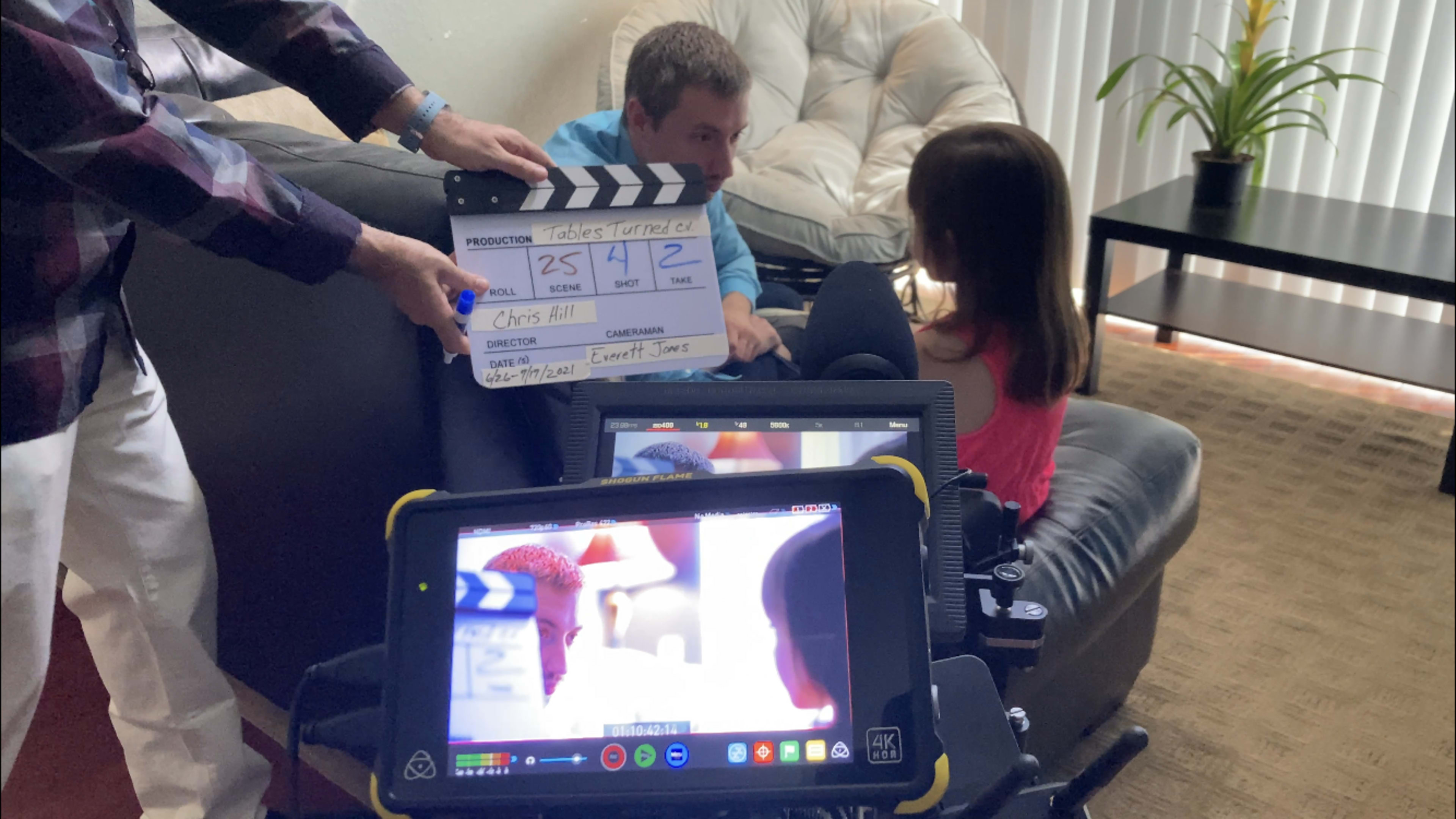 A young boy and a girl sit on a couch while a person holds a film clapperboard in front of them during a video shoot.