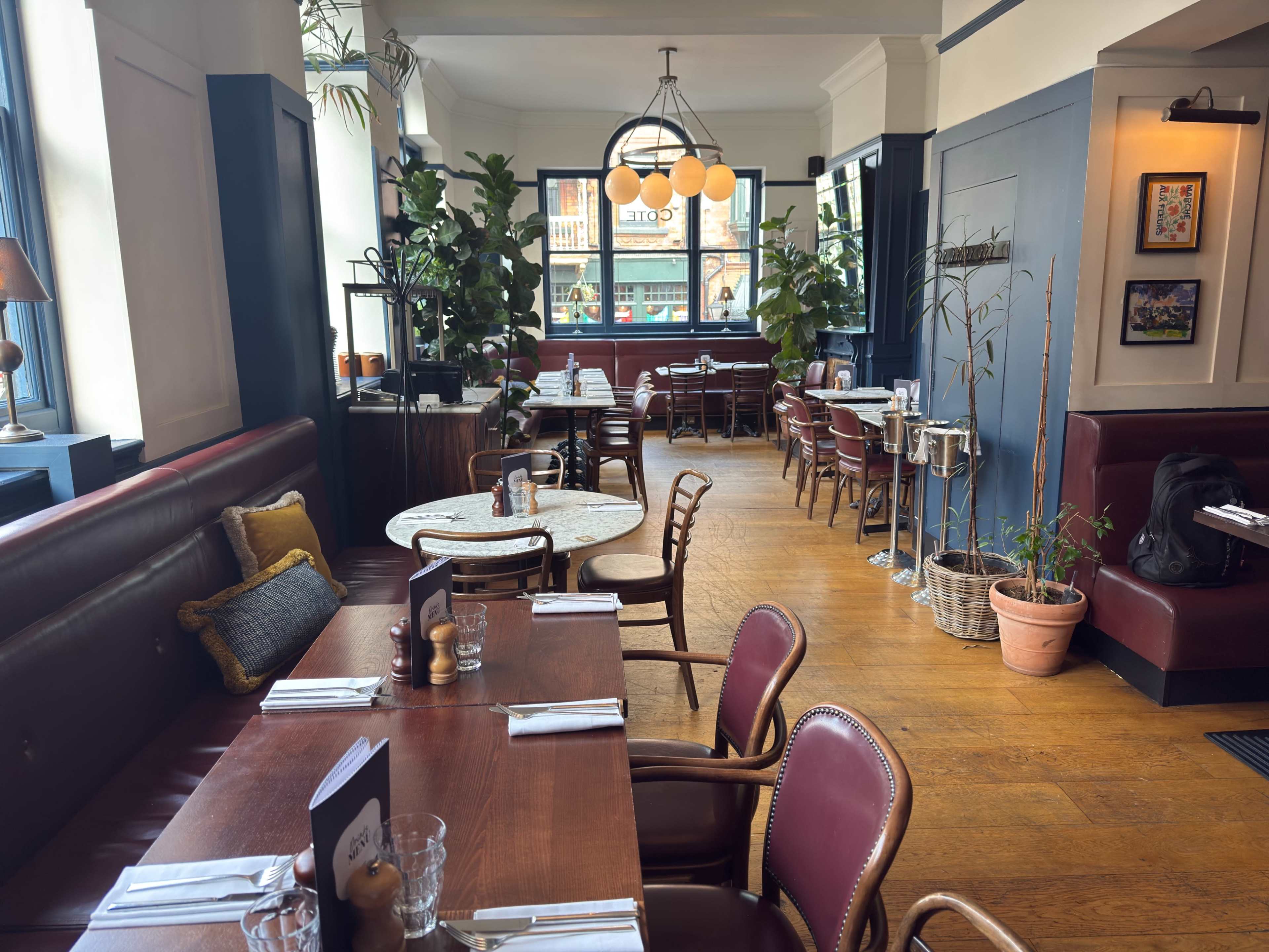 The image shows the interior of a restaurant featuring wooden tables, leather seating, and potted plants with large windows allowing natural light to fill the space.